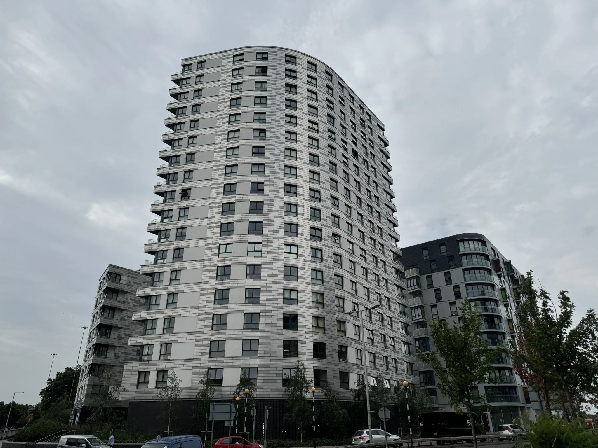 Tall modern high-rise residential building with curved design and multiple balconies, seen under a cloudy sky.