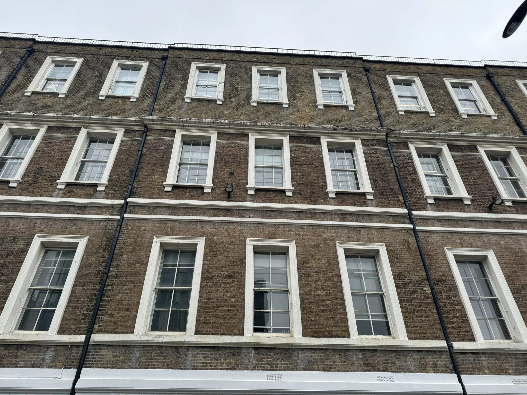 View of a brick building with multiple white-framed windows, black external pipes, and a white trim near the bottom, under a cloudy sky.
