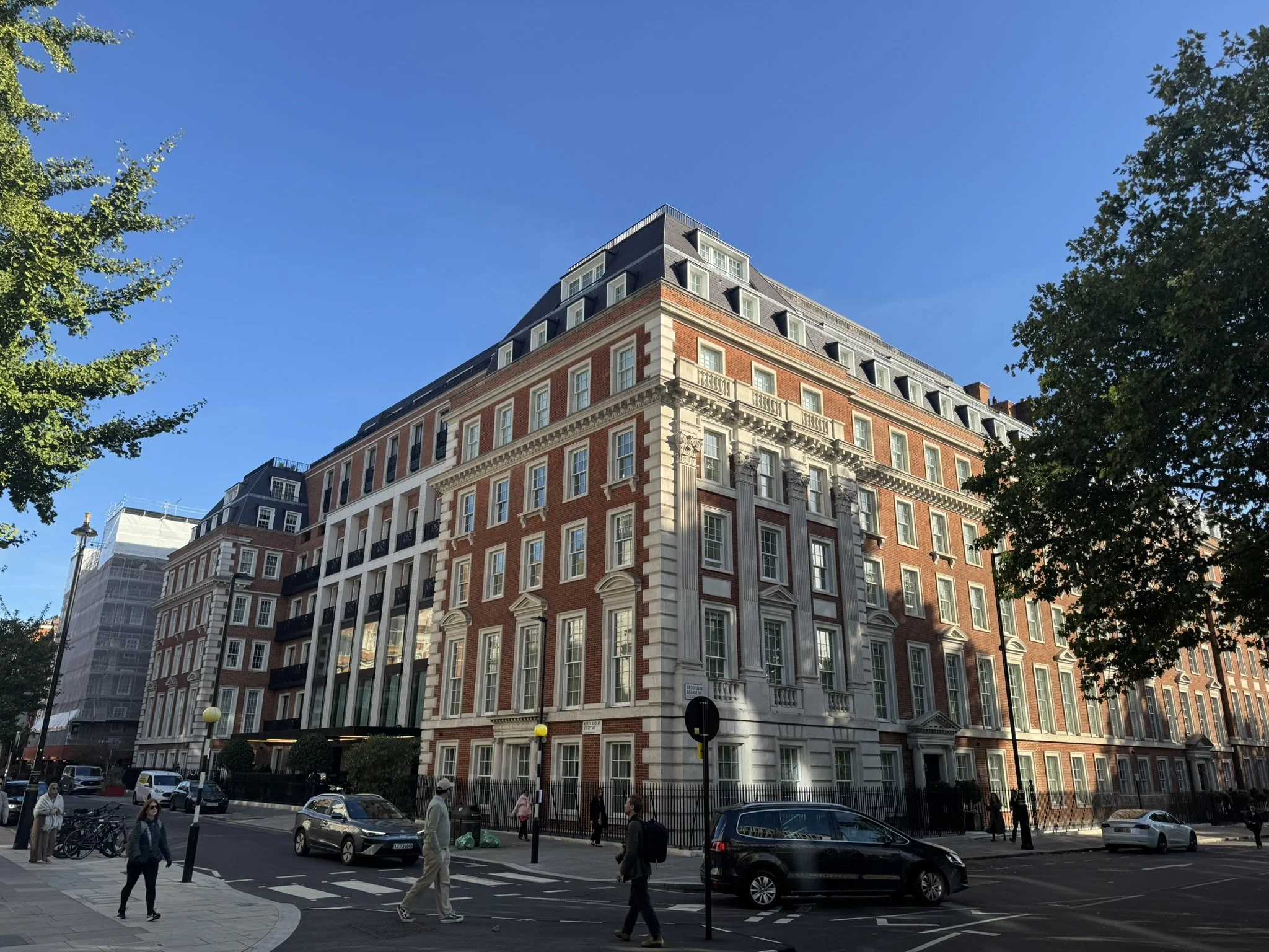 A large brick and stone corner building with multiple floors, rows of windows, and decorative architectural details in an urban setting with pedestrians and cars on the street, and trees framing the scene, under a clear blue sky.