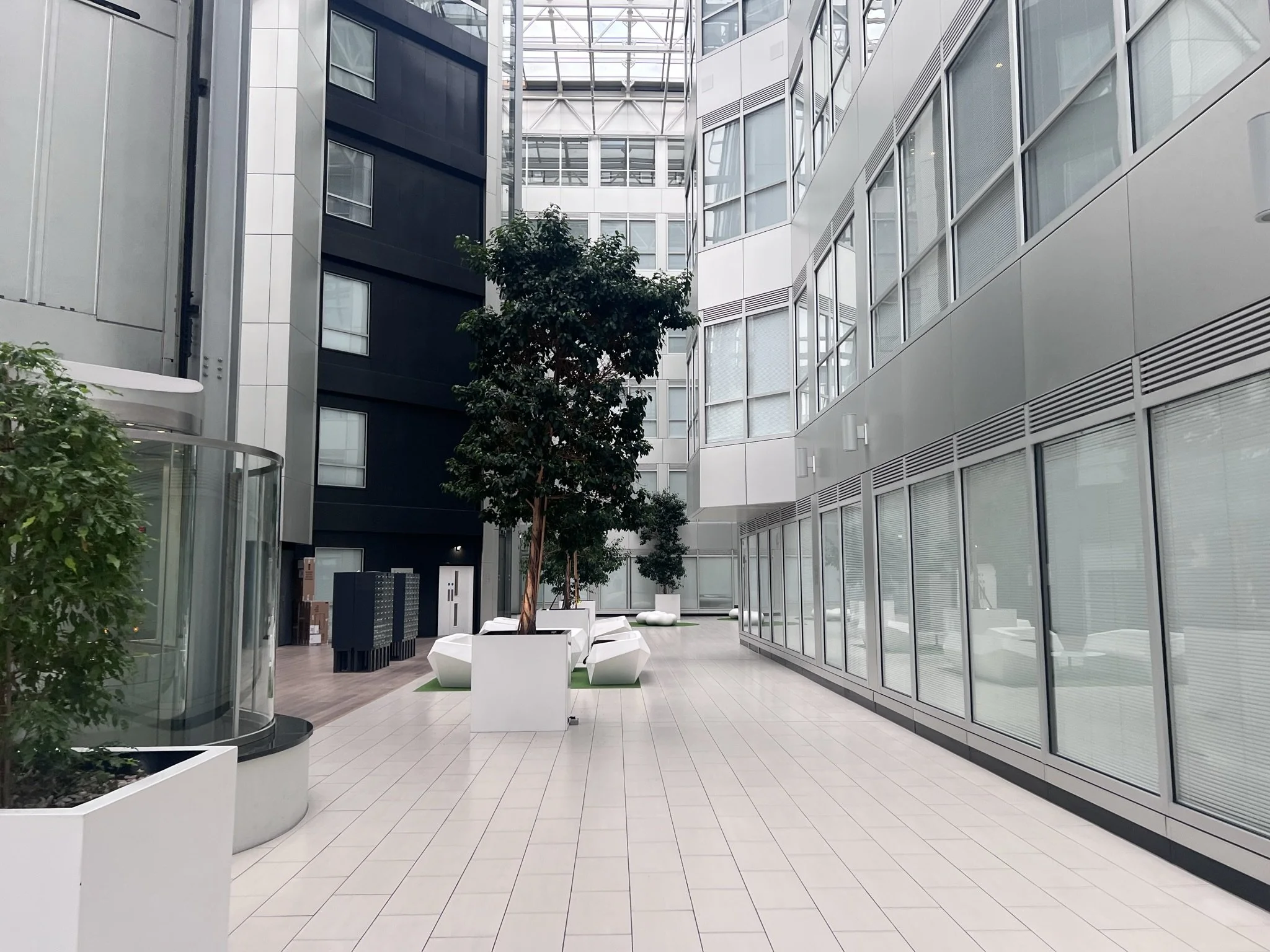 Modern indoor atrium or common area with white tiled flooring, glass walls, and multiple trees in white planters, surrounded by high-rise buildings.