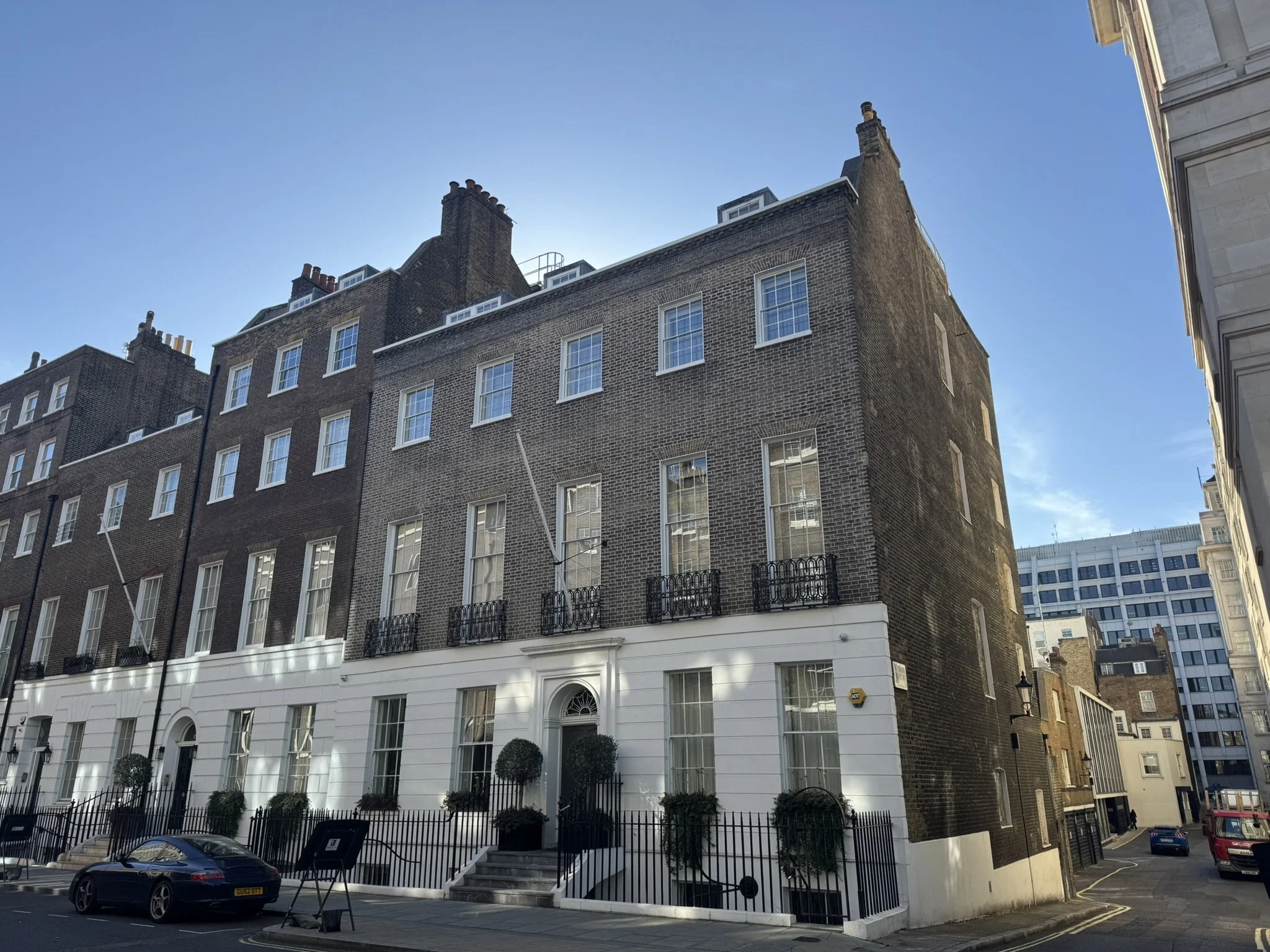 Street view of a multi-story brick building with white trim, windows with black wrought-iron balconies, small doors with white frames, and potted bushes outside. Modern buildings are visible in the background under a clear blue sky.