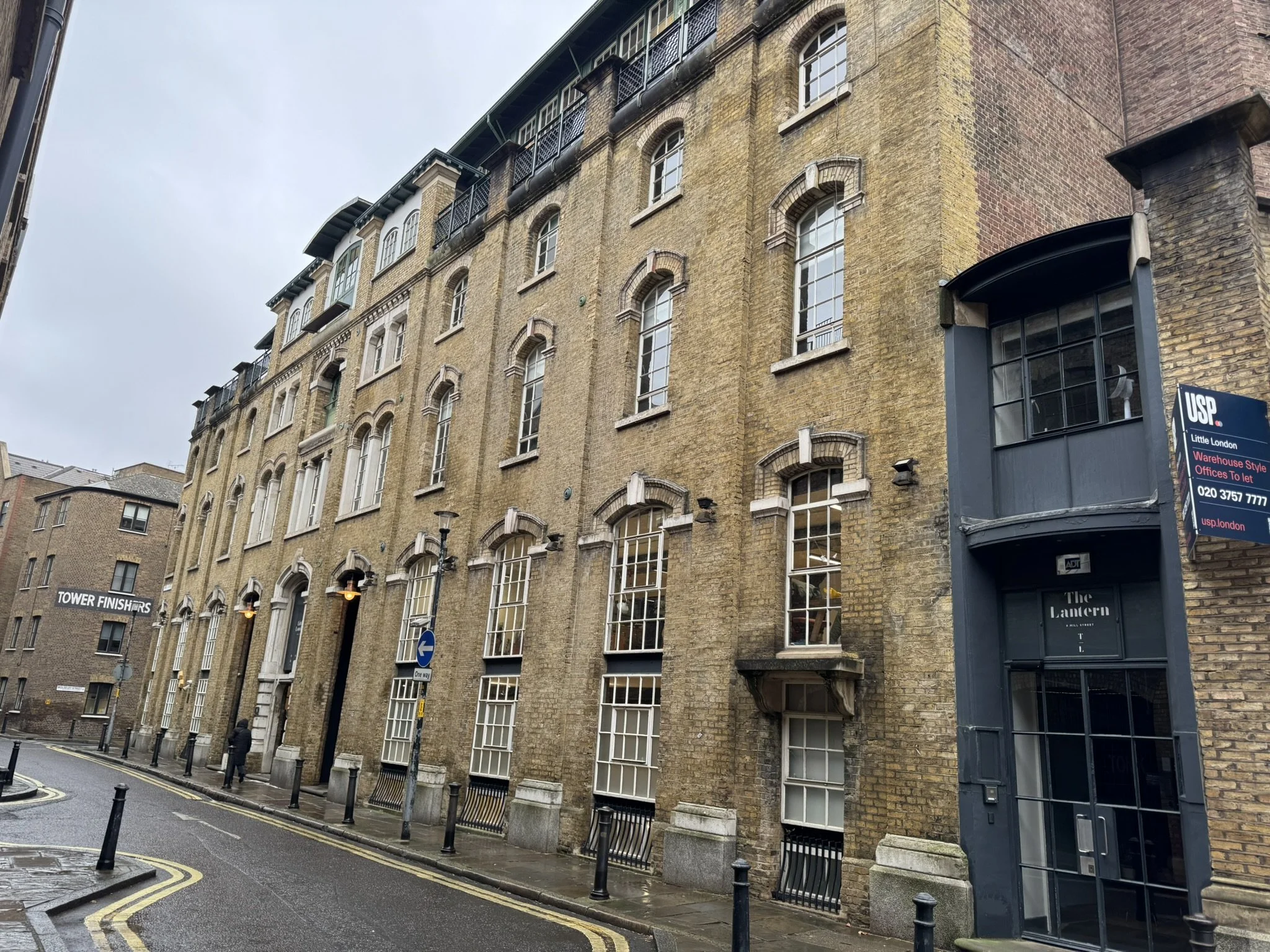 A row of multi-story brick buildings on a rainy street with street lamps, bollards, and a man walking. The building has large windows and an entrance with a sign reading 'The Lantern.'