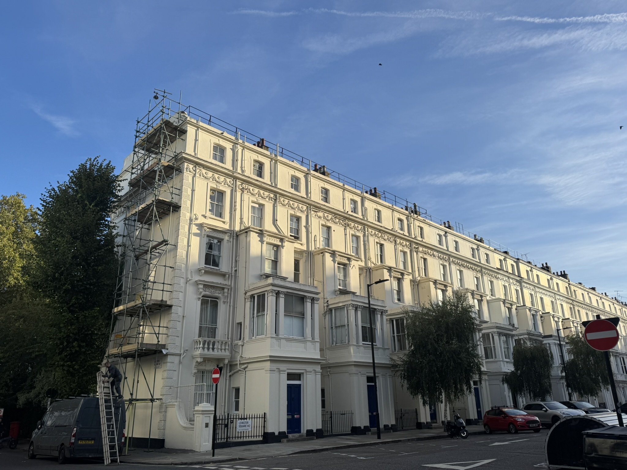 A large cream-colored multi-story building with ornate architectural details, surrounded by trees, parked cars, and a street. Scaffolding is visible on the left side of the building, and there are clear blue skies overhead.