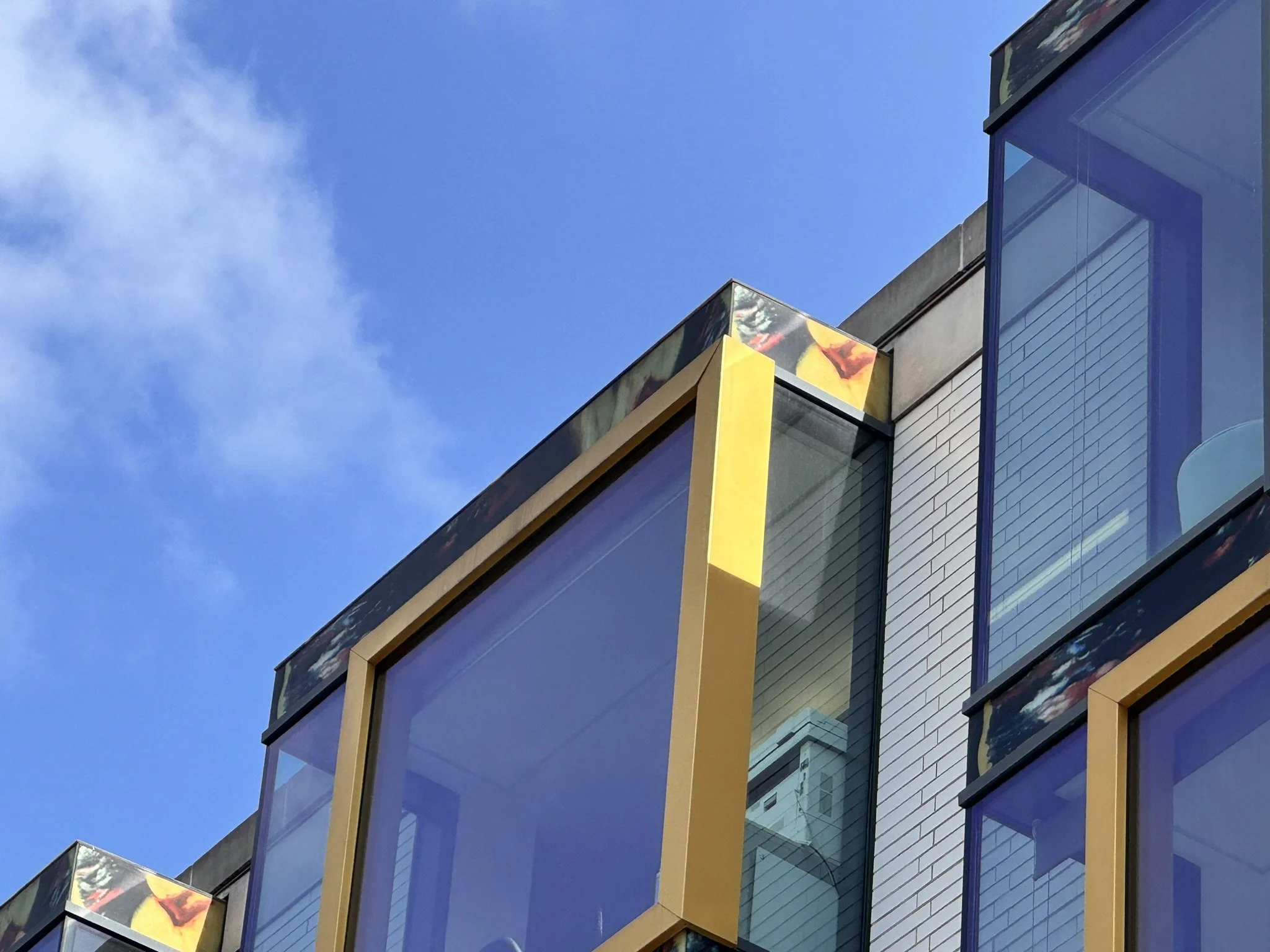 Close-up view of modern building windows with yellow framing and a part of the building's brick exterior; clear blue sky with some clouds in the background.