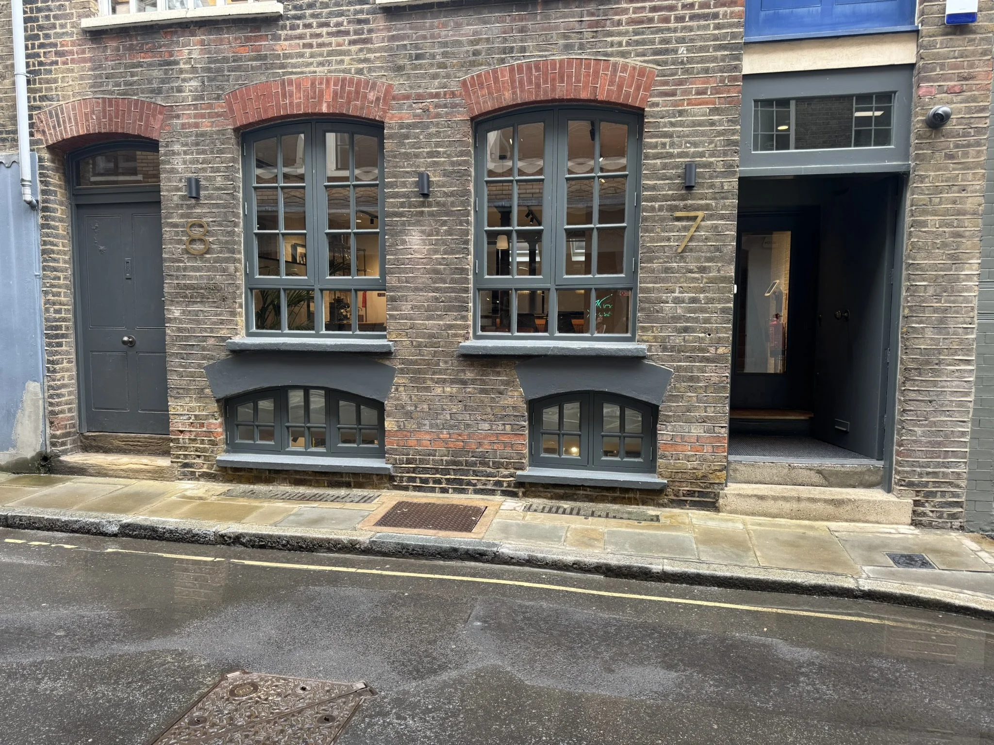 Two brick buildings on a rain-soaked street, each with gray doors and windows. The left building has the number 8, the right building has the number 7. The buildings feature large rectangular and smaller arched windows with gray frames.