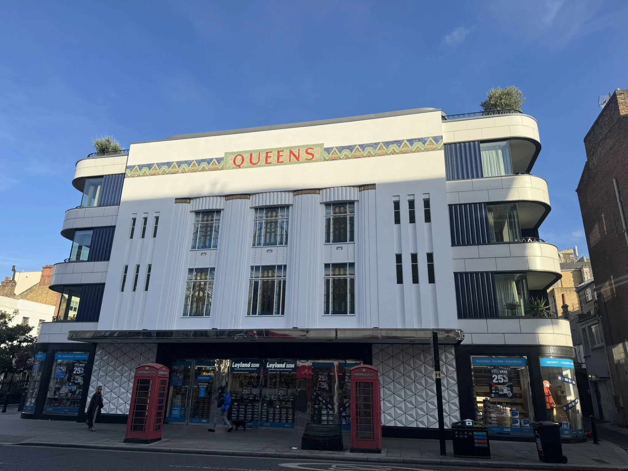 A modern white multi-story building with rounded balconies and large windows, with a sign that reads 'QUEENS' at the top. The ground floor appears to be a shop with glass windows and doors, advertising sales and products, and there are people walking on the sidewalk in front.