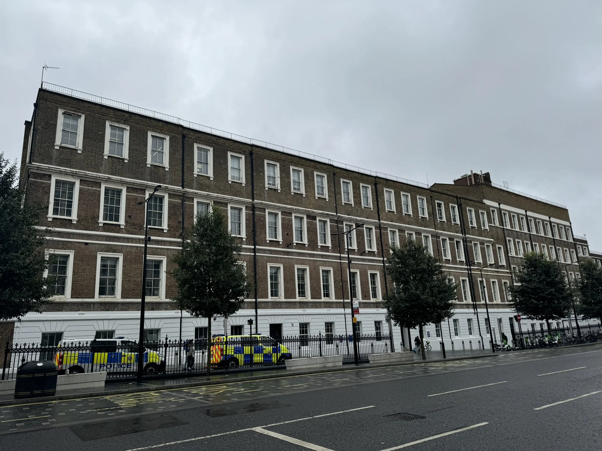 A multi-story brick building with numerous windows, trees lining the sidewalk, and police vehicles parked along the street on a cloudy day.