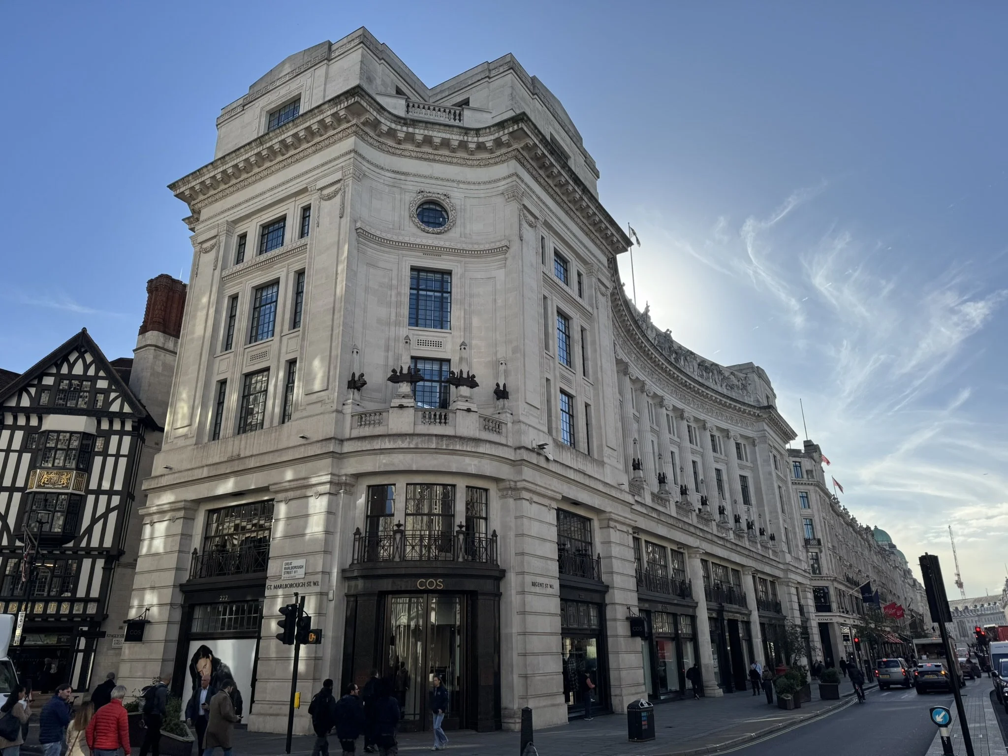 Street view of a curved historic building with ornate architecture, large windows, and retail shops on the ground floor, with pedestrians and cars in front.