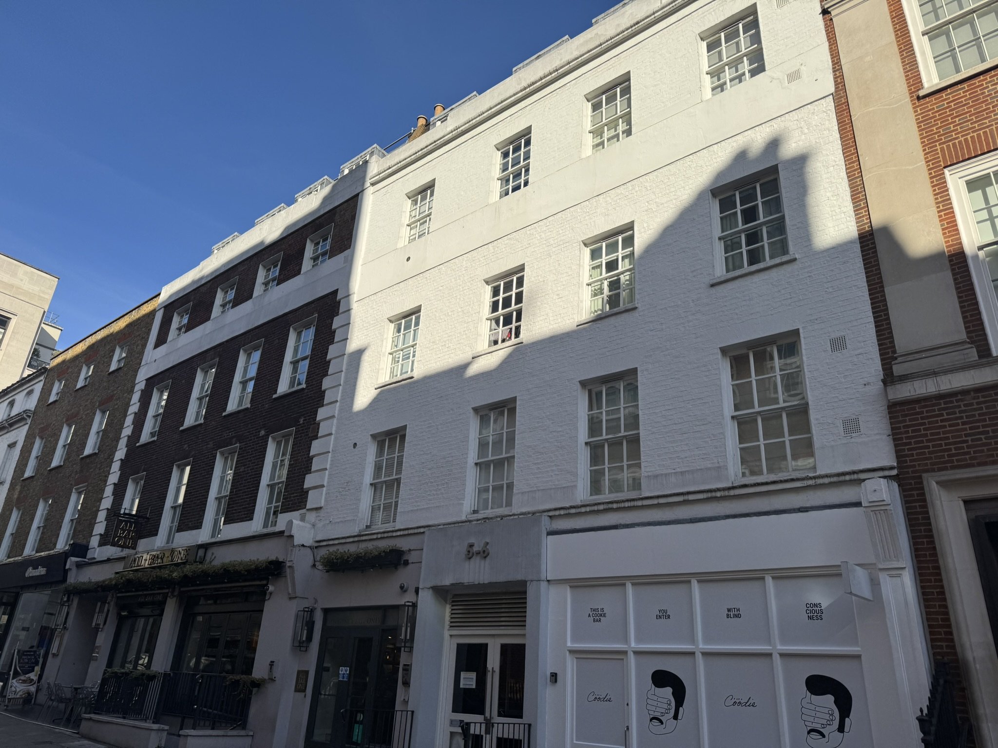 A city street with a four-story building that has a white facade and multiple windows, with a shadow of an adjacent building cast onto it under a clear blue sky.