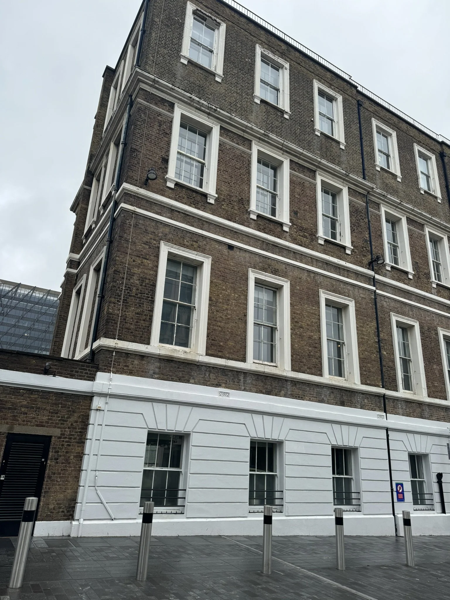 A multi-story brick building with white window frames and a white base, on a street with bollards and a parking sign, under an overcast sky.