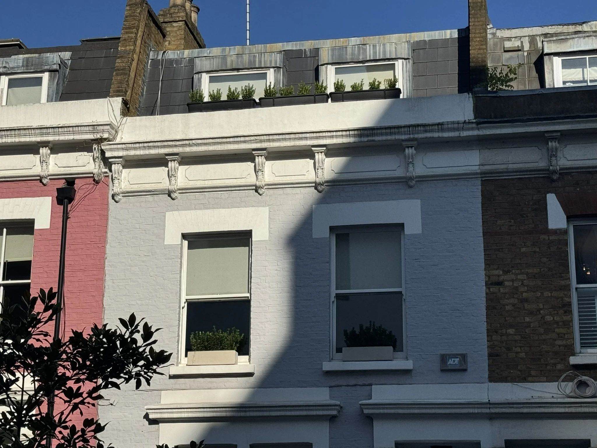 Exterior view of a row of colorful townhouses, focusing on a white building with three windows and decorative architectural details, with a shadow cast over part of the facade, and neighboring buildings partially visible.