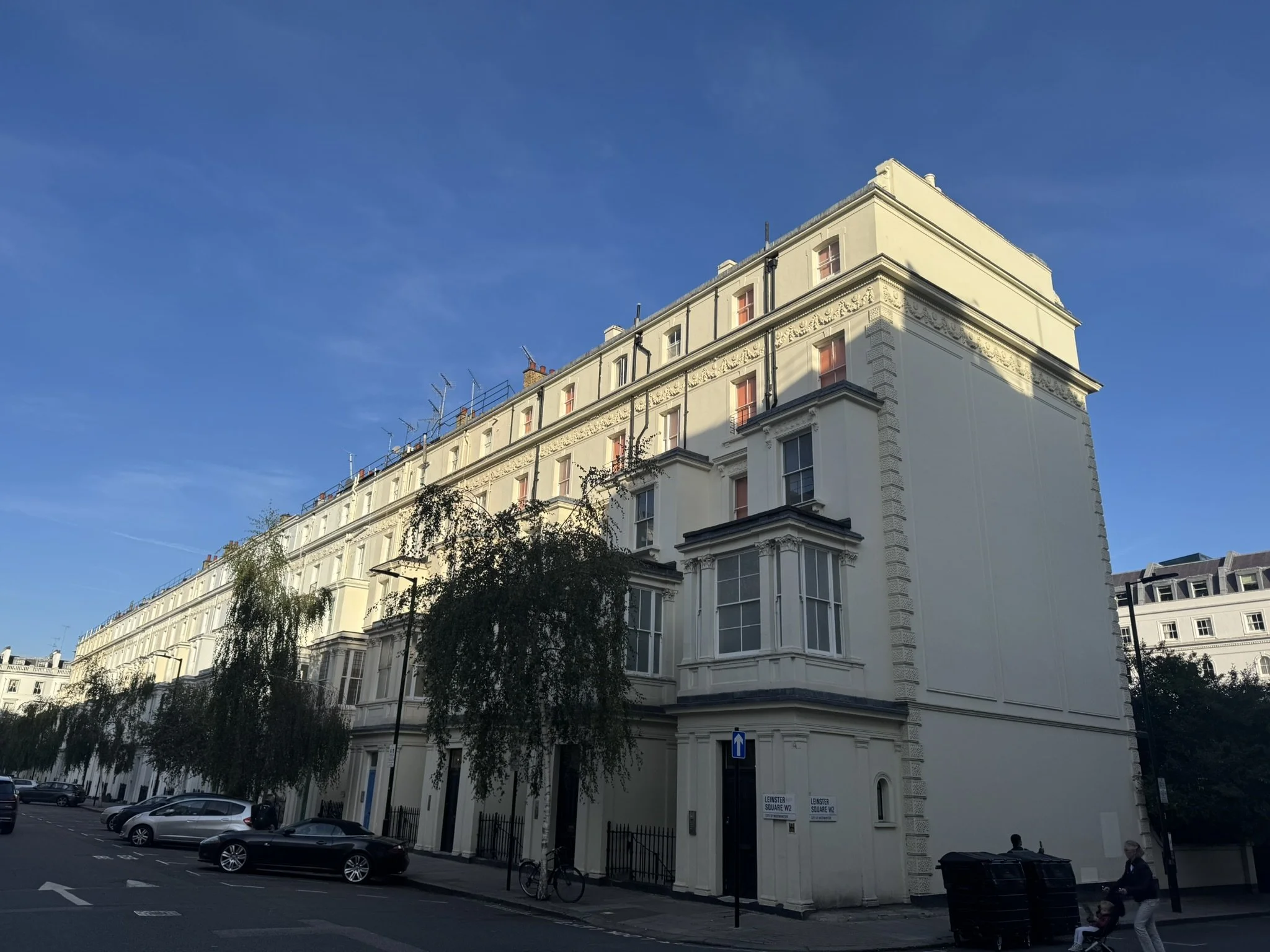 A cream-colored, multi-story Victorian-style building on a city street with blue sky overhead. There are parked cars and a few pedestrians, including a person pushing a stroller.