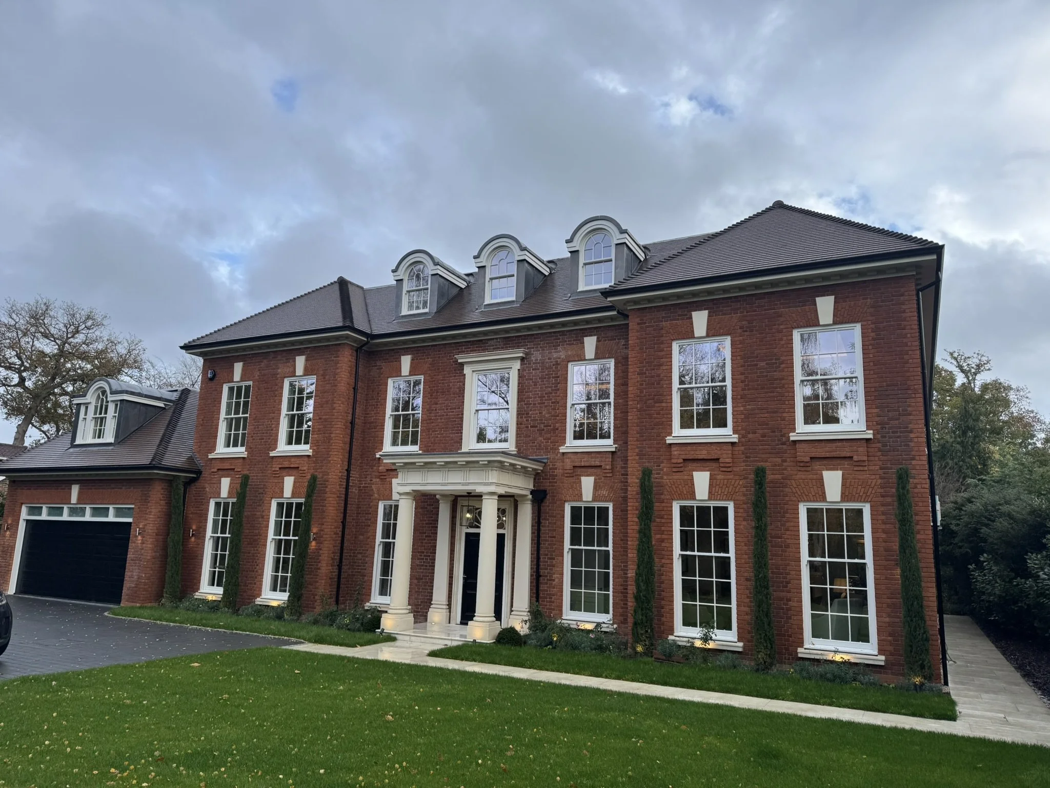 A large, three-story brick house with a dark shingled roof, white trim, multiple windows, and a covered front porch with columns. There is a driveway on the left side and a well-maintained green lawn in front.