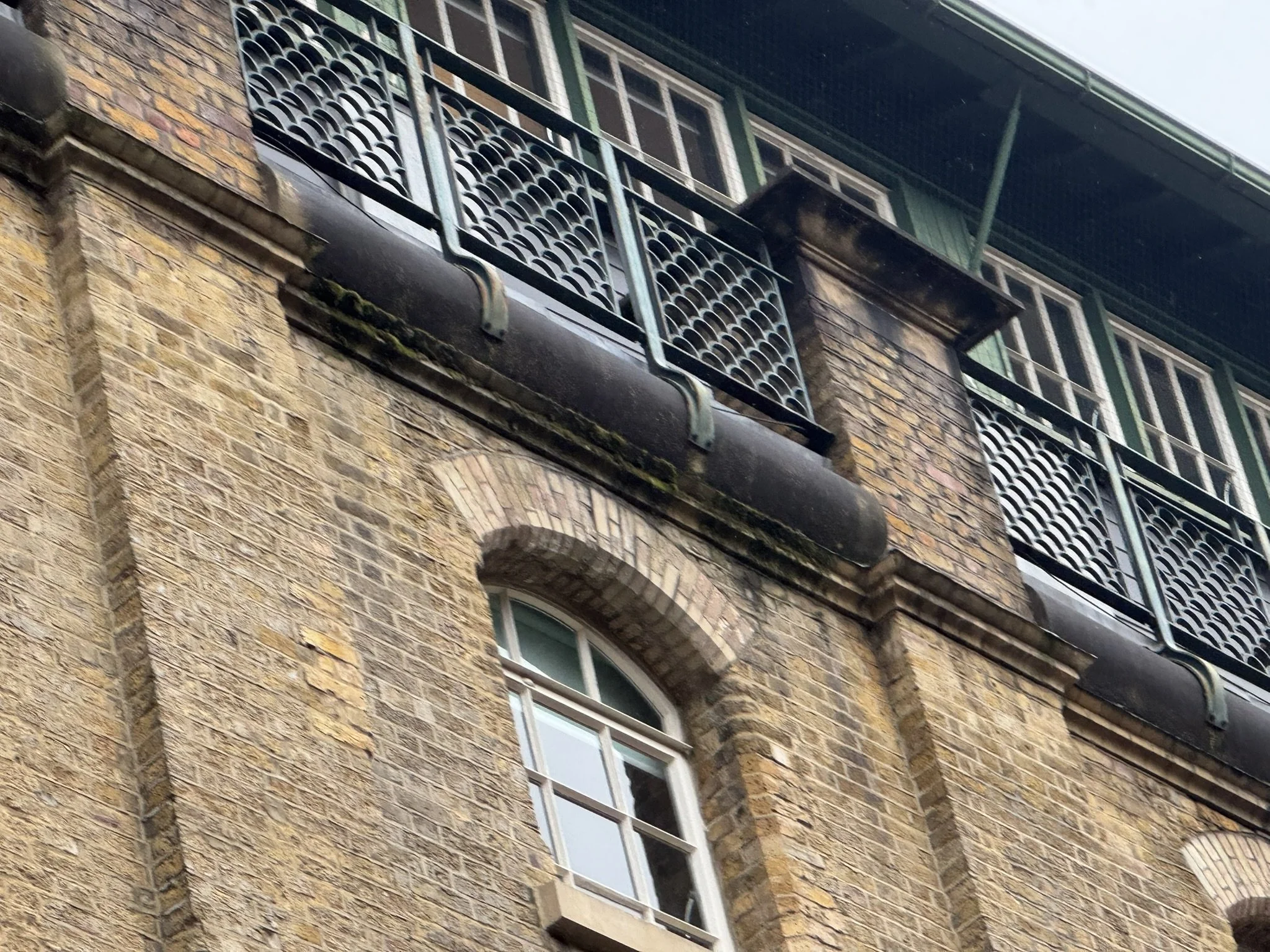 Close-up of an old brick building with a single small window and a metal balcony with decorative railings on the upper floor.