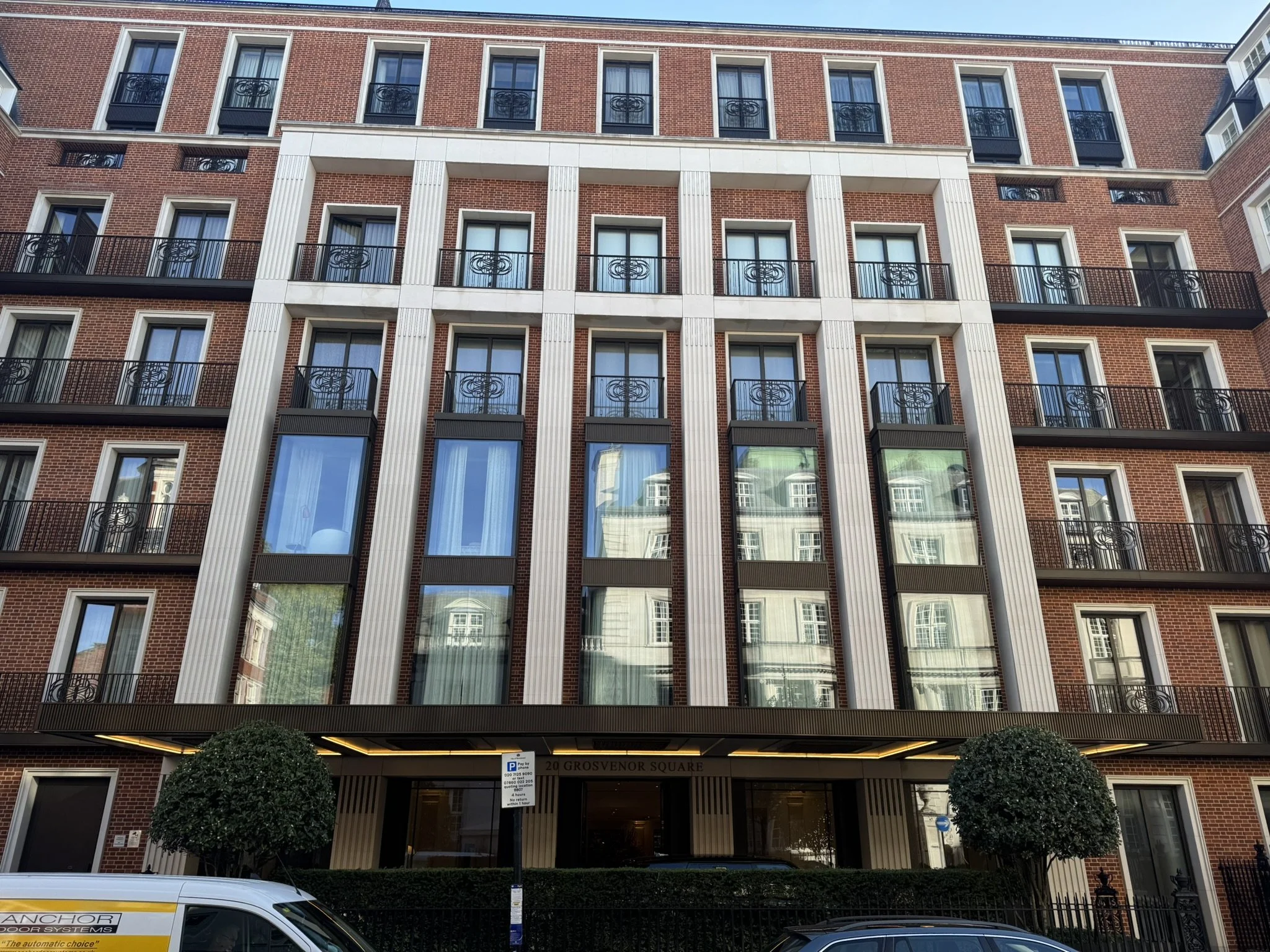 Front view of a multi-story brick building with several windows and balconies, reflected in large glass windows, with trees and parked cars in front.