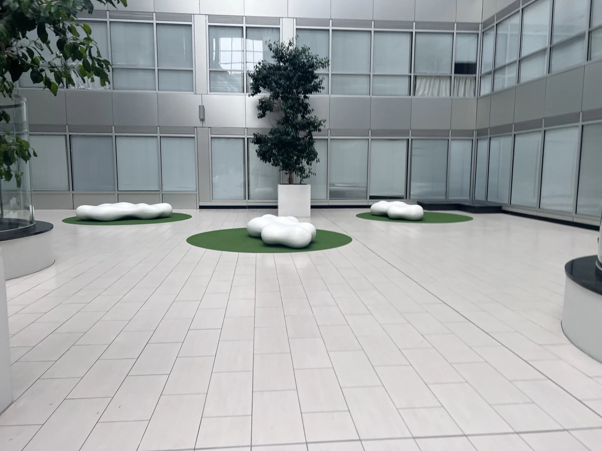 Indoor courtyard with white tiled floor, large glass windows on walls, and modern white cloud-shaped seating areas on green rugs surrounding a potted tree in the center.
