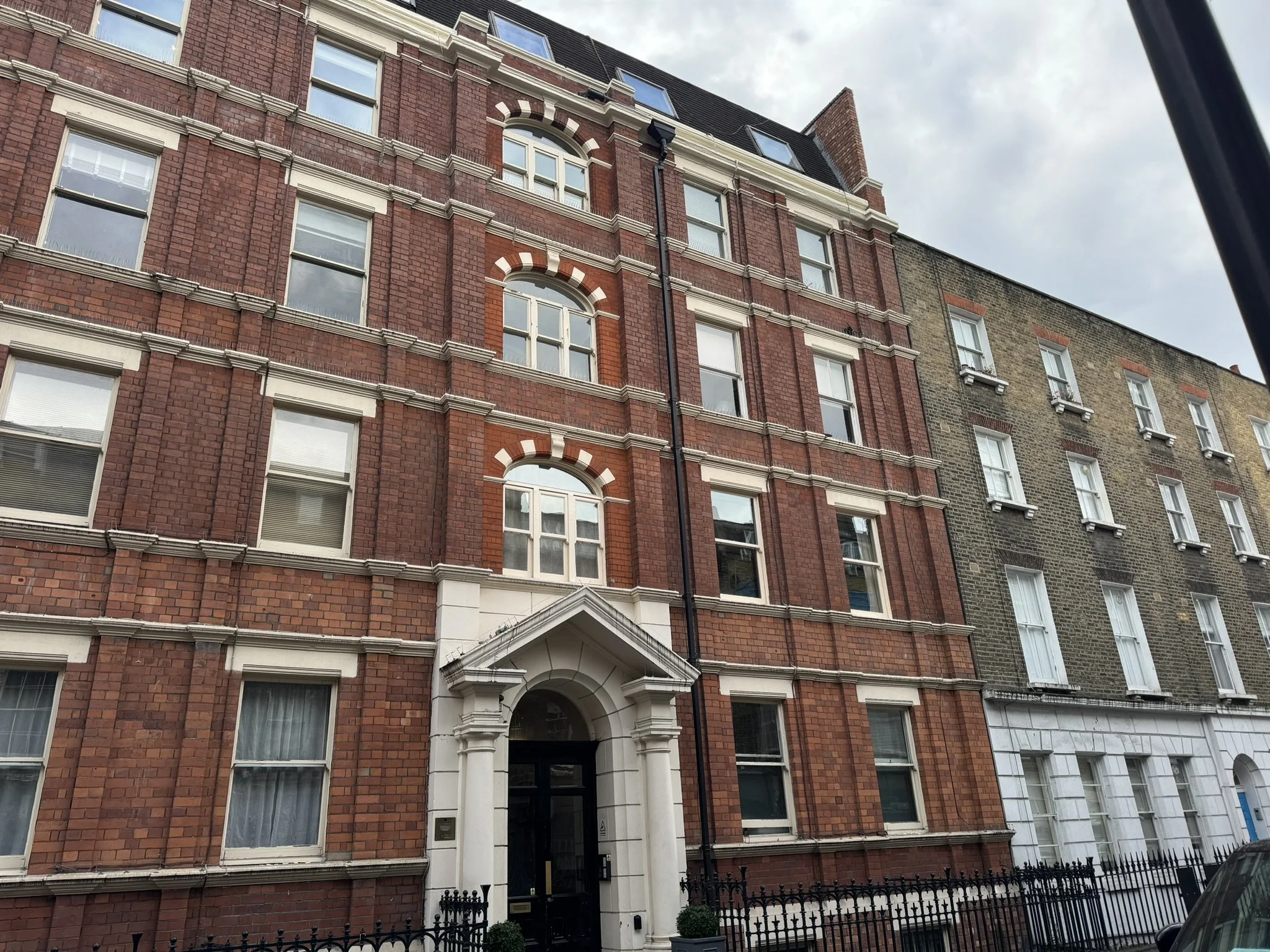 A multi-story brick building with arched windows and white decorative trim, featuring a prominent entryway with an arched doorway and columns. The building is next to other similar structures, with a black metal fence in front.