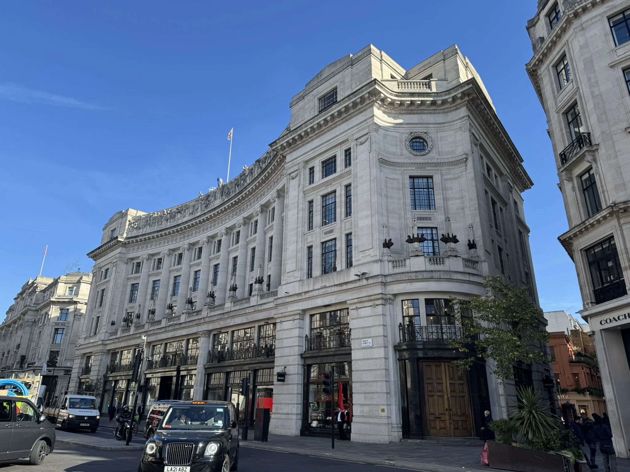 A large historic building with ornate architecture on a busy city street, with cars, buses, and pedestrians in front, under a clear blue sky.