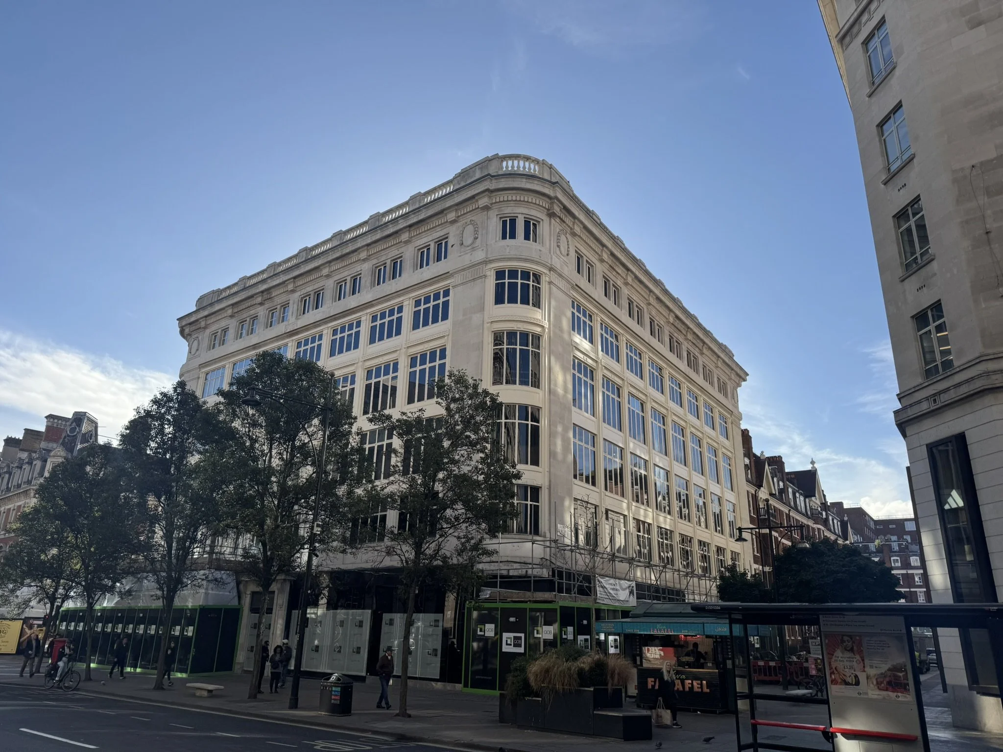 A large multi-story white building with large windows, on a city street with trees, people, and a bus stop.