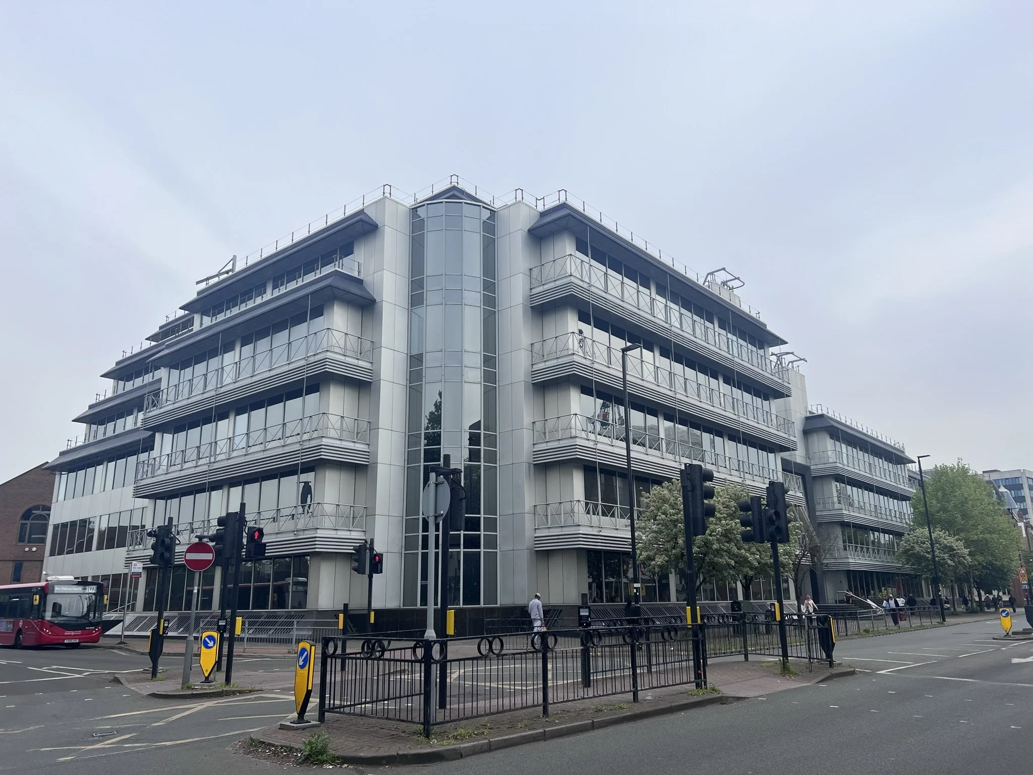 Modern multi-story office building with glass and metal exterior, balconies on each floor, at a city intersection with traffic lights and a pink bus nearby.