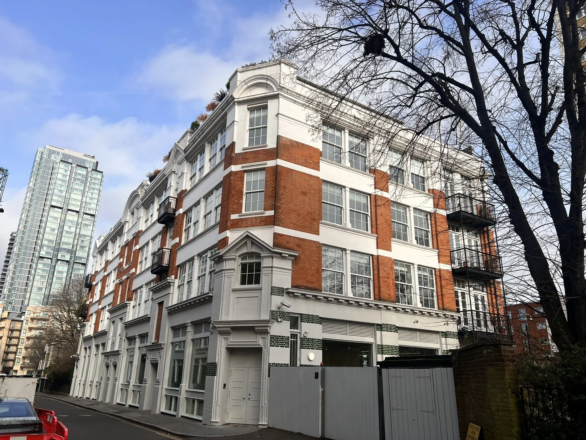 A multi-story brick and white building with bay windows and small balconies on the upper floors, located on a corner street with leafless trees and a modern high-rise building in the background.