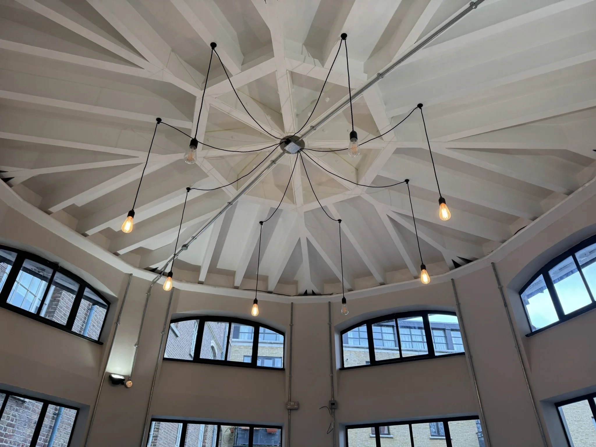 Ceiling with white geometric beams and hanging Edison-style light bulbs in a round room with large arched windows.