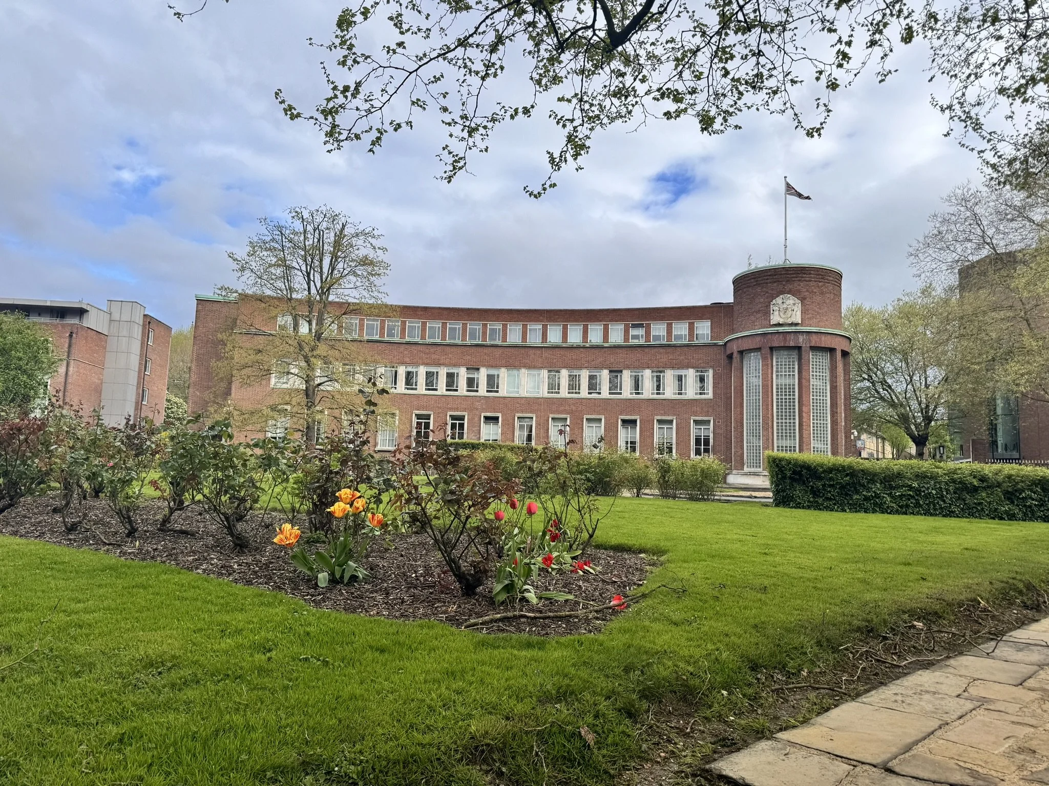 A brick building with a curved tower and flag on top, surrounded by a well-manicured lawn and flower bed with tulips. Trees with budding leaves and a cloudy sky in the background.