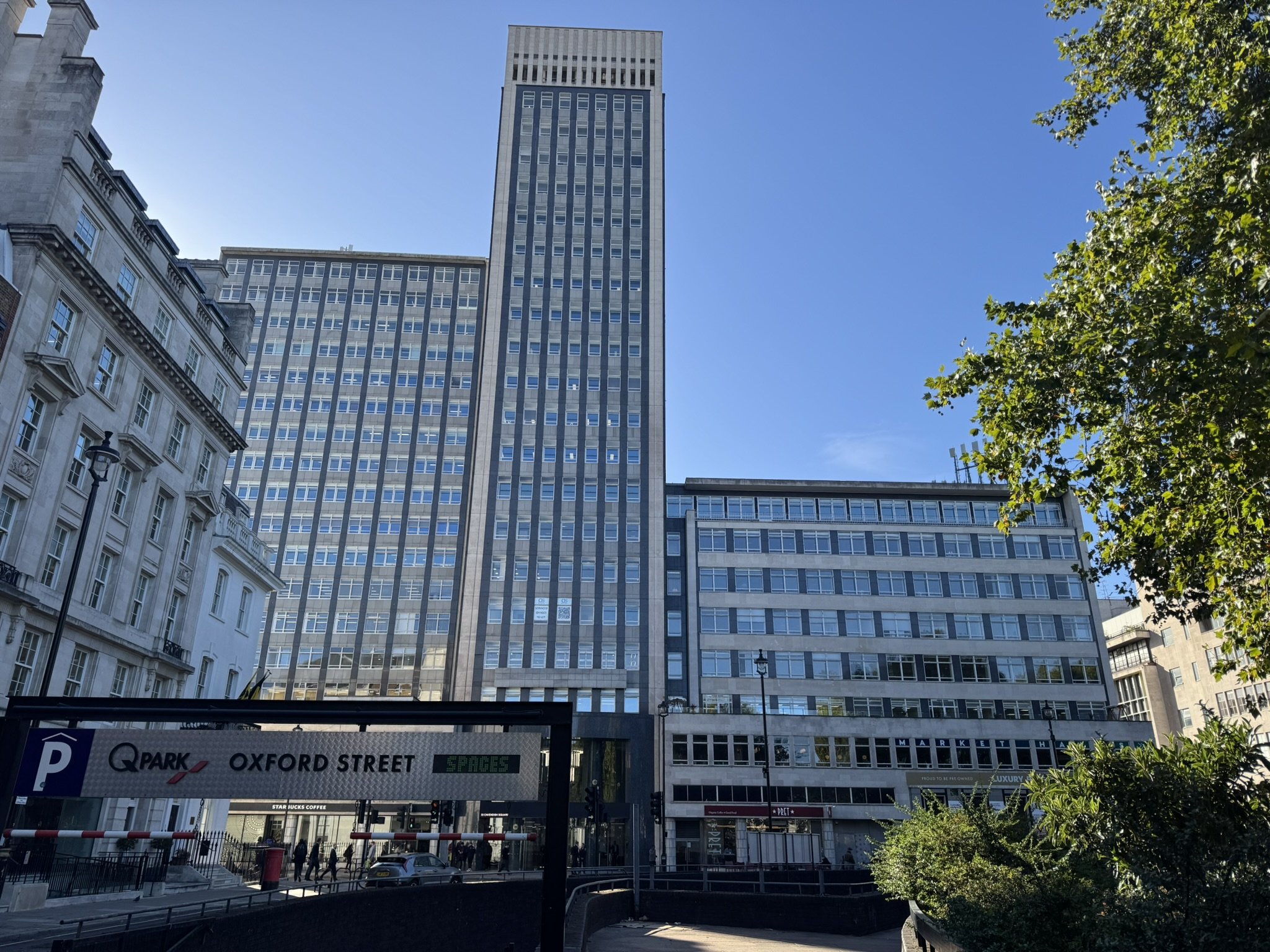 Tall modern office building in an urban area with other buildings and trees, parking sign visible in foreground, clear blue sky.