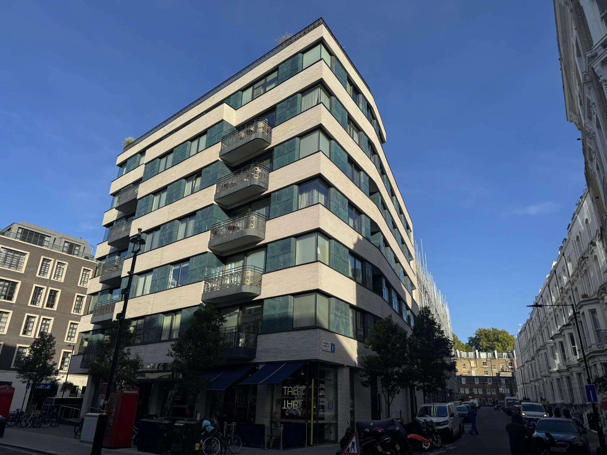 A modern multi-story building with glass windows and curved balconies, standing on a city street during daytime with blue sky and nearby white buildings.