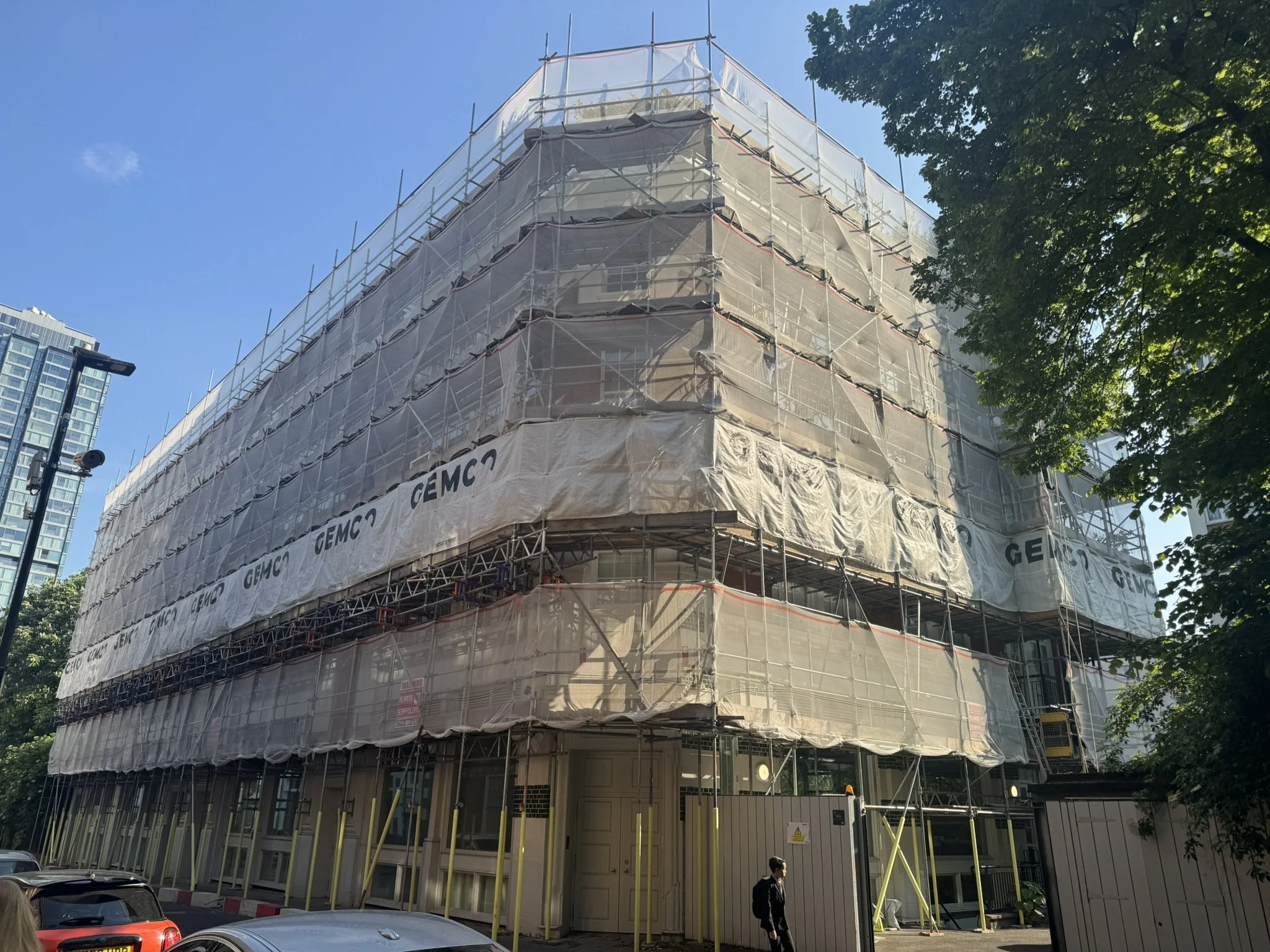 Building under construction with scaffolding and white protective sheeting, city street scene with cars and a person walking.