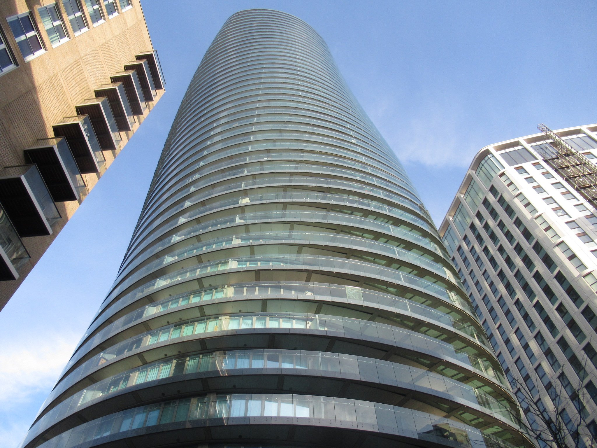 Low-angle view of a modern glass skyscraper surrounded by other tall buildings against a blue sky.
