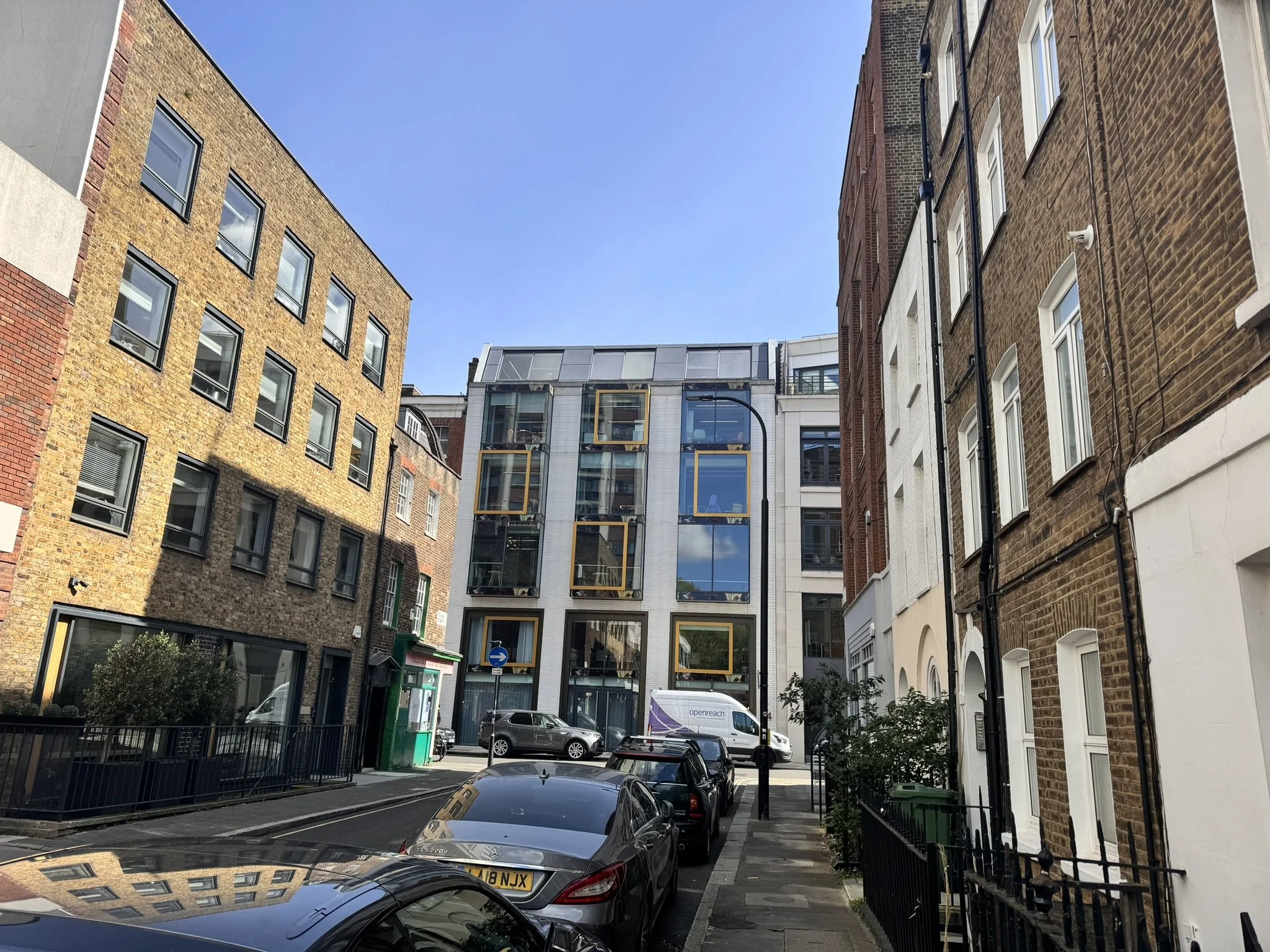 City street view with parked cars, modern multi-story buildings, and a clear blue sky.