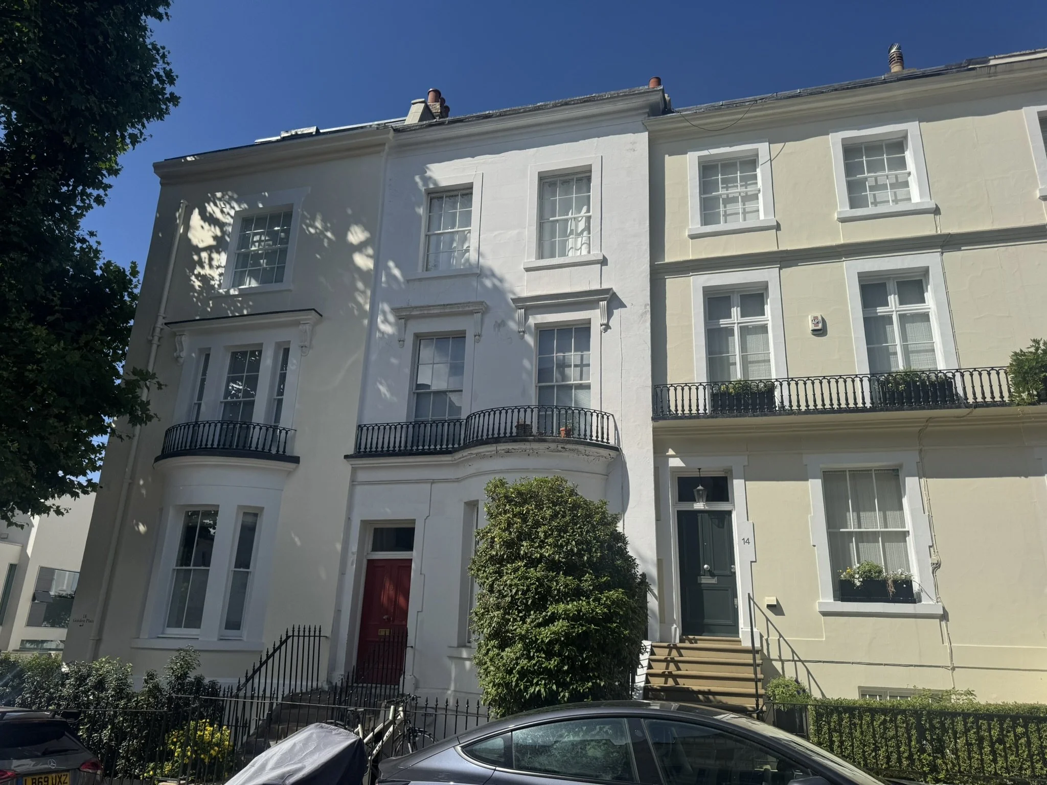Three-story white and cream-colored residential building with black railing balconies, a green bush in front, parked motorcycle and car, blue sky, and tree shadows.