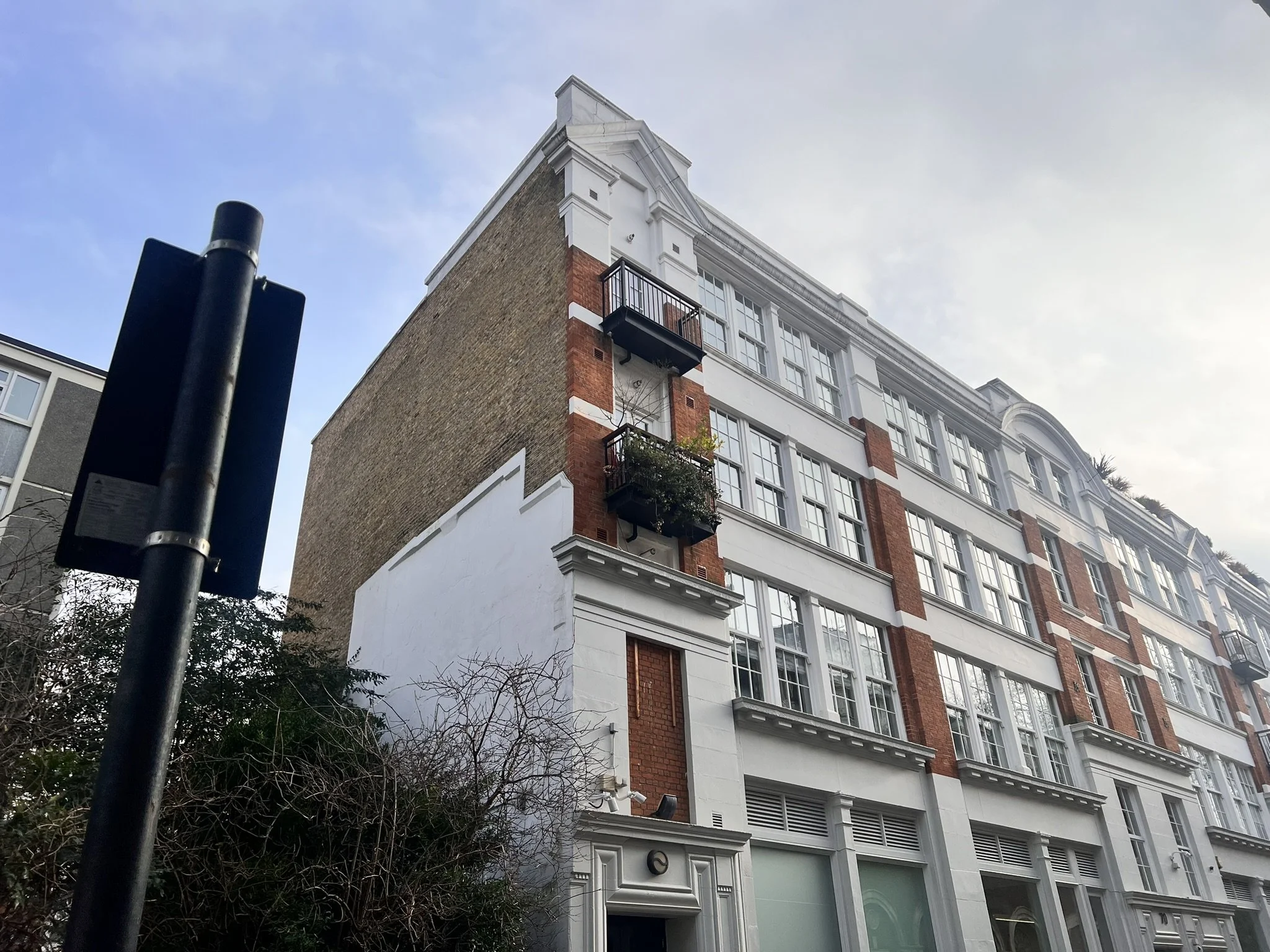 A multi-story building with white and red brick exterior, multiple windows, and small balconies with plants, under a partly cloudy sky.