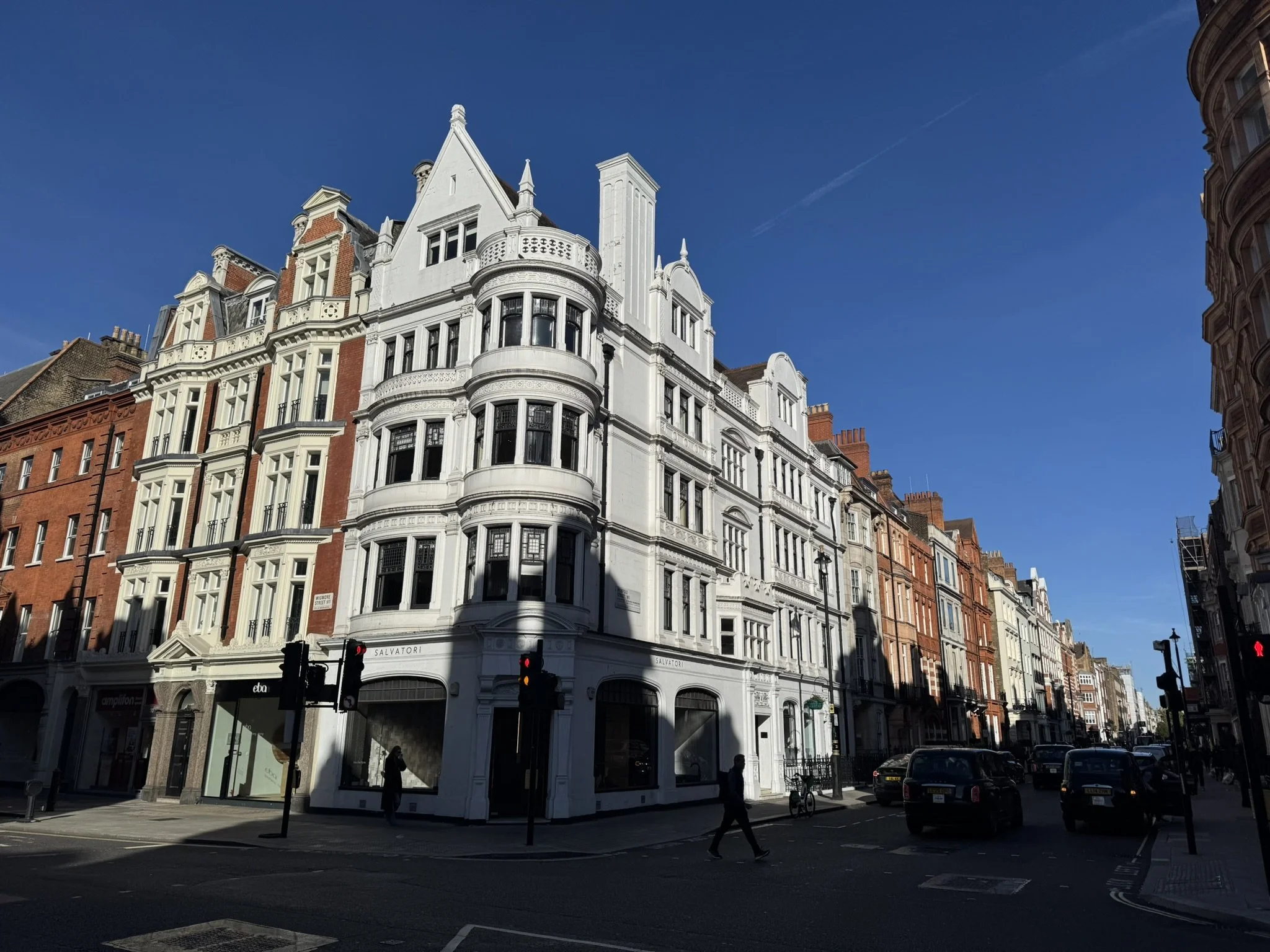 Street view of a white, Victorian-style building on a corner, with shops at the ground level and residential or office space above, under a clear blue sky, with a few pedestrians and cars at the intersection.
