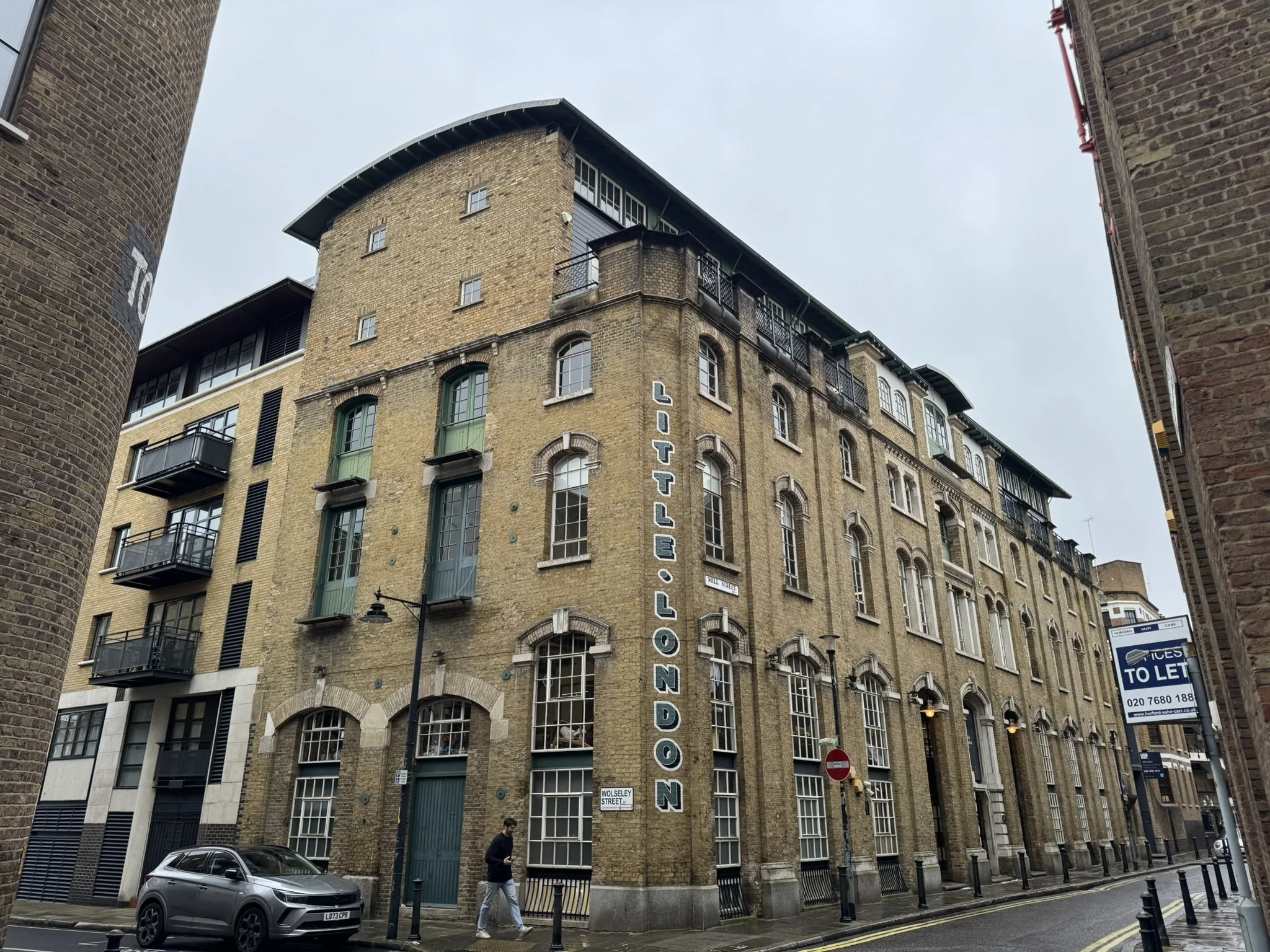 A large historic brick building with a vertical sign reading 'LITTLE LONDON' on the corner of a street in London, England. The building has multiple windows, some with small balconies, and street lamps outside. There are cars parked and a person walking on the sidewalk.