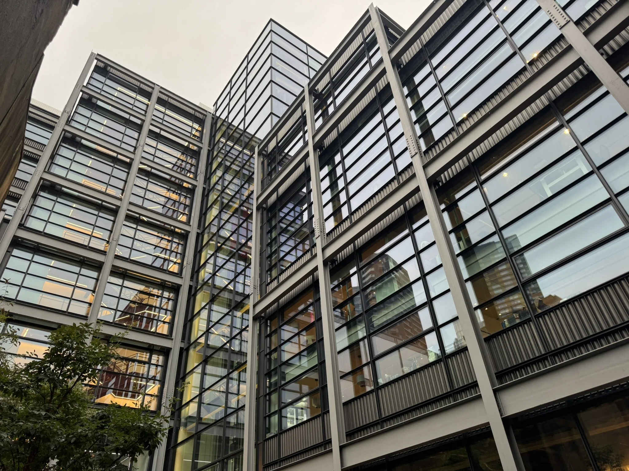 Modern glass office building with multiple floors and reflections of neighboring structures, trees visible in the foreground, and an overcast sky.