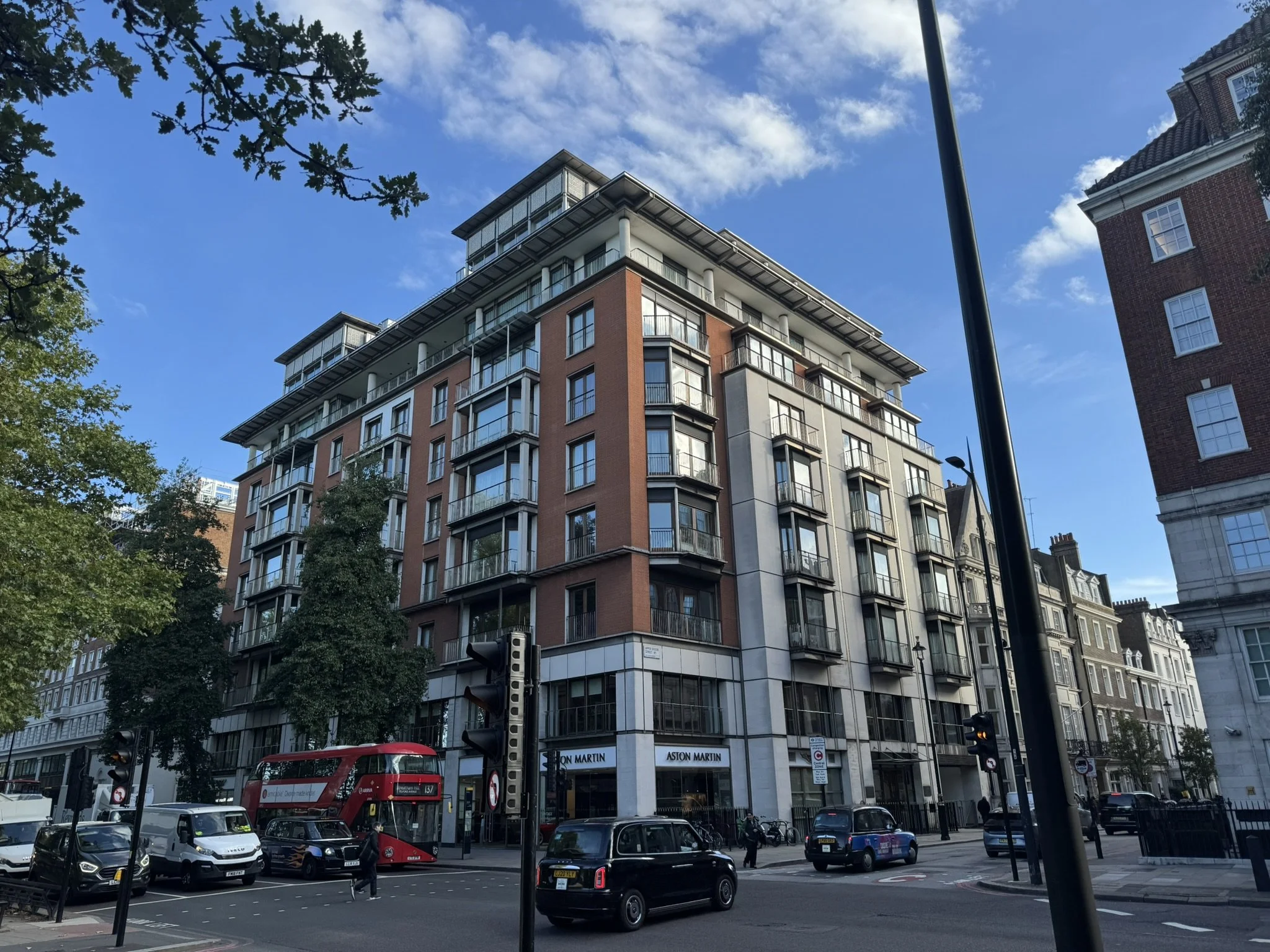 Urban street scene with a modern multi-story building, a red double-decker bus, and cars, under a partly cloudy sky during daytime.