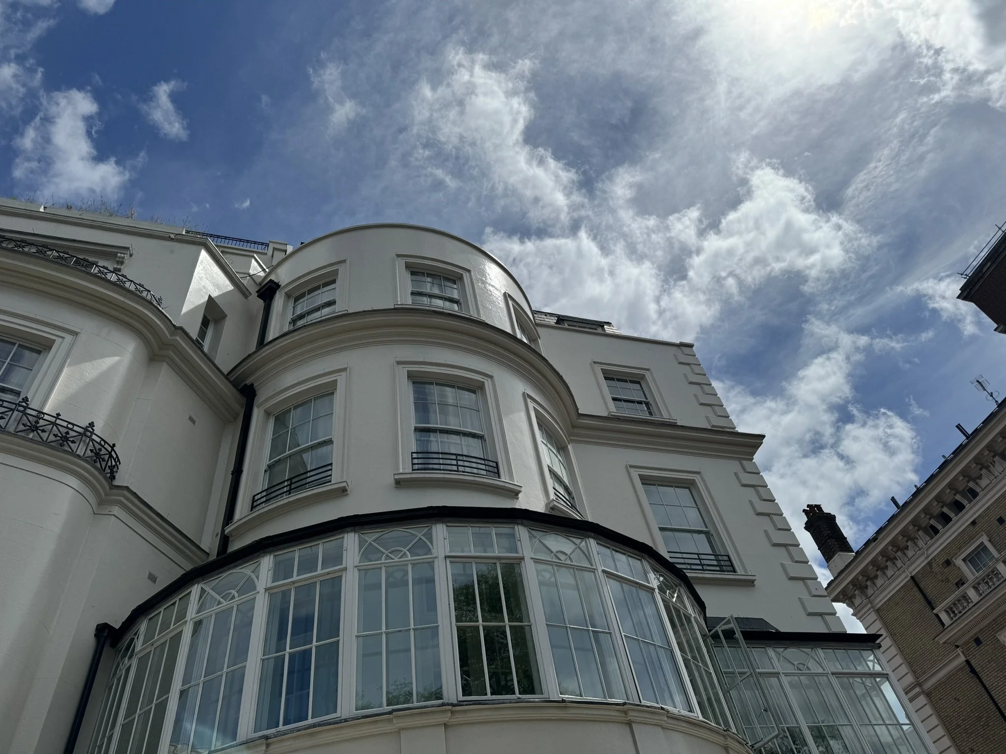 Low-angle view of a white, multi-story building with curved balconies and large windows, against a partly cloudy sky.