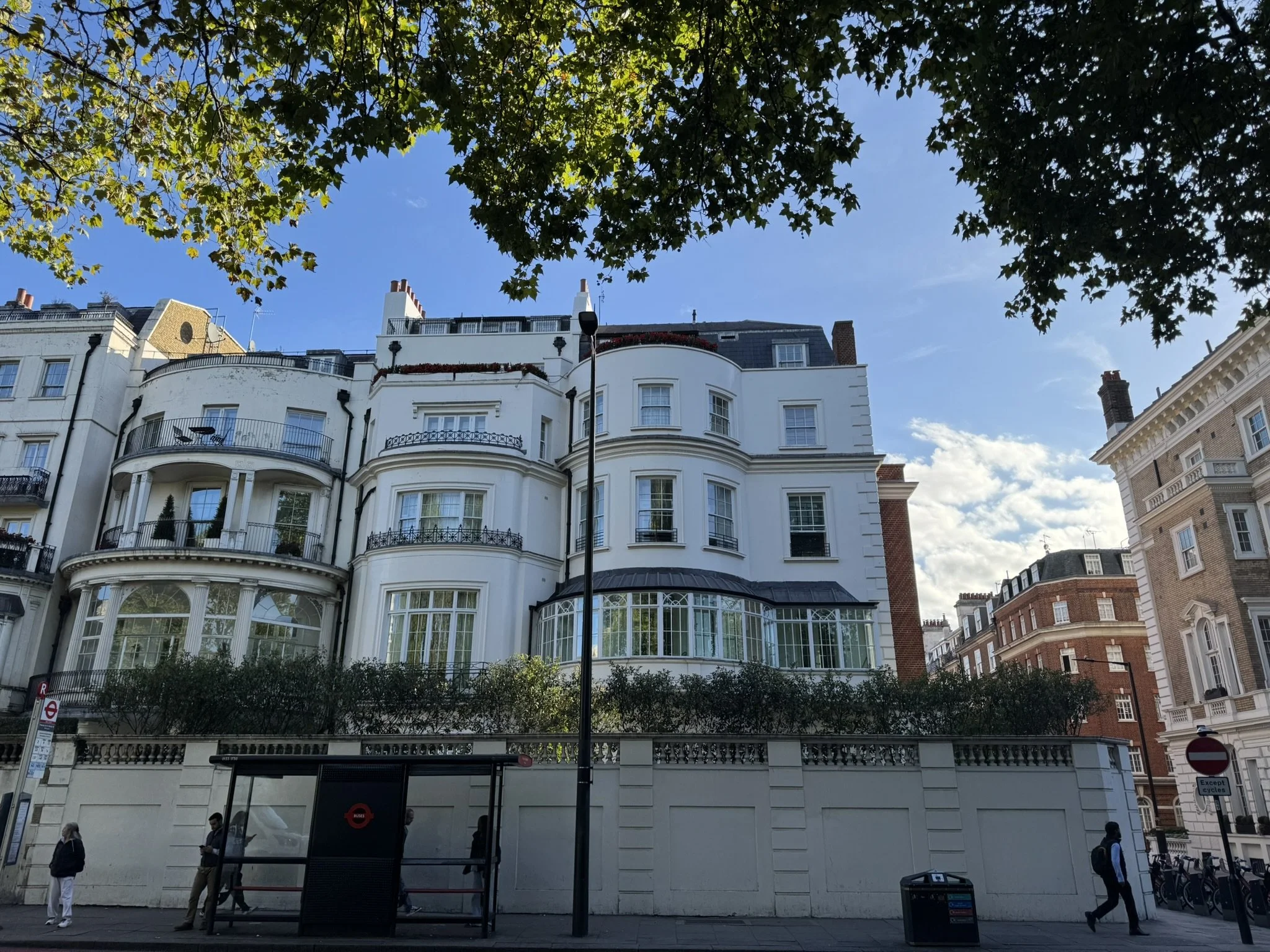 Street view with a white multi-story building with rounded balconies and large windows, surrounded by trees, under a partly cloudy sky.