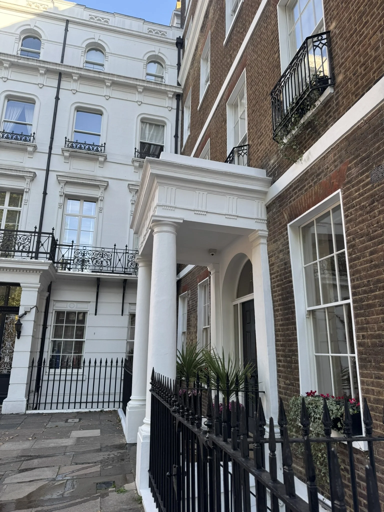 Close-up view of the entrance to a townhouse with black wrought iron fence, white columns, and a small garden with potted plants