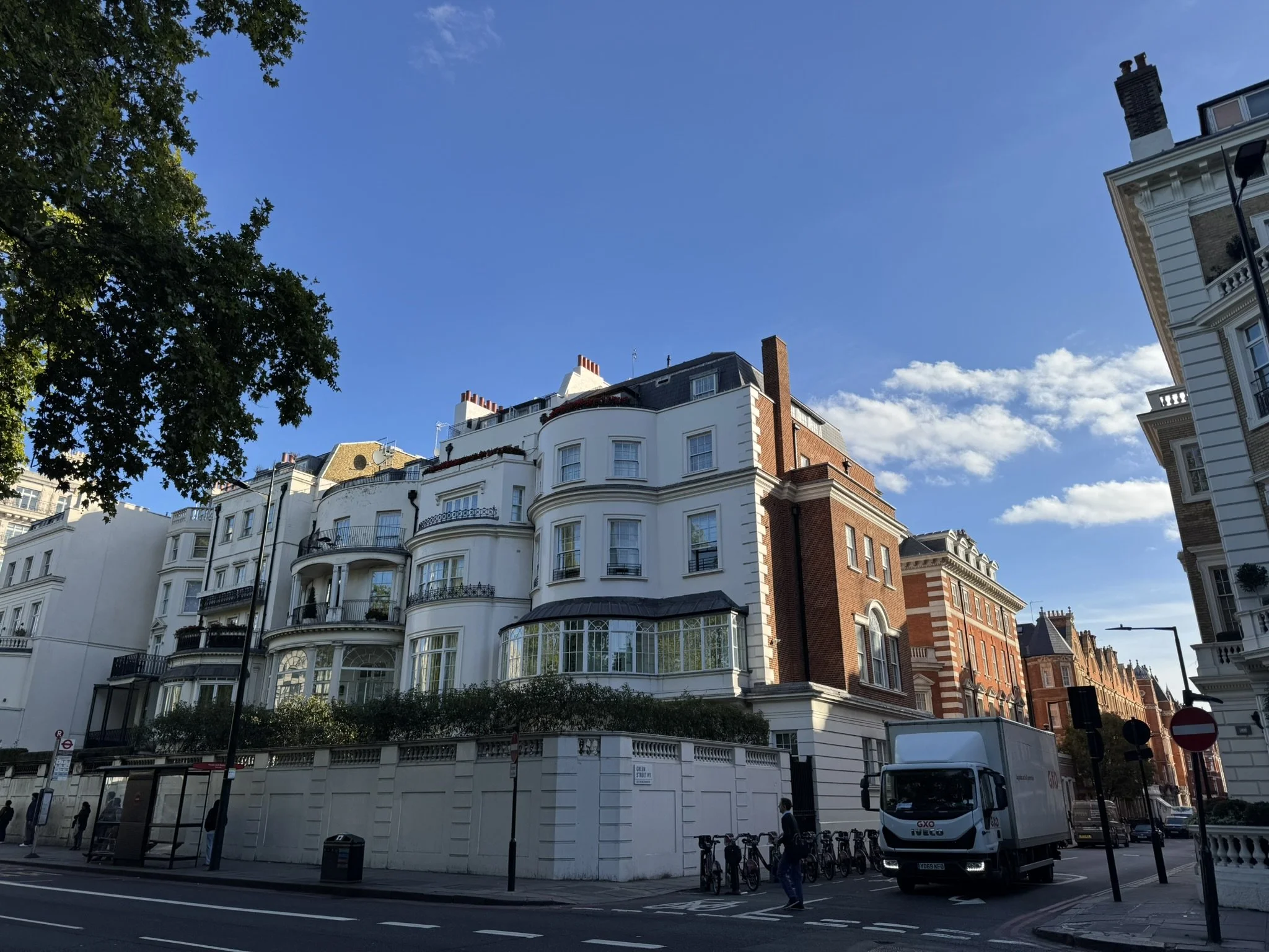Street view of a white and red brick multi-story building with curved balconies, large windows, and a black roof under a blue sky with some clouds. There are trees, bicycles, a white delivery truck, and pedestrians on the sidewalk.