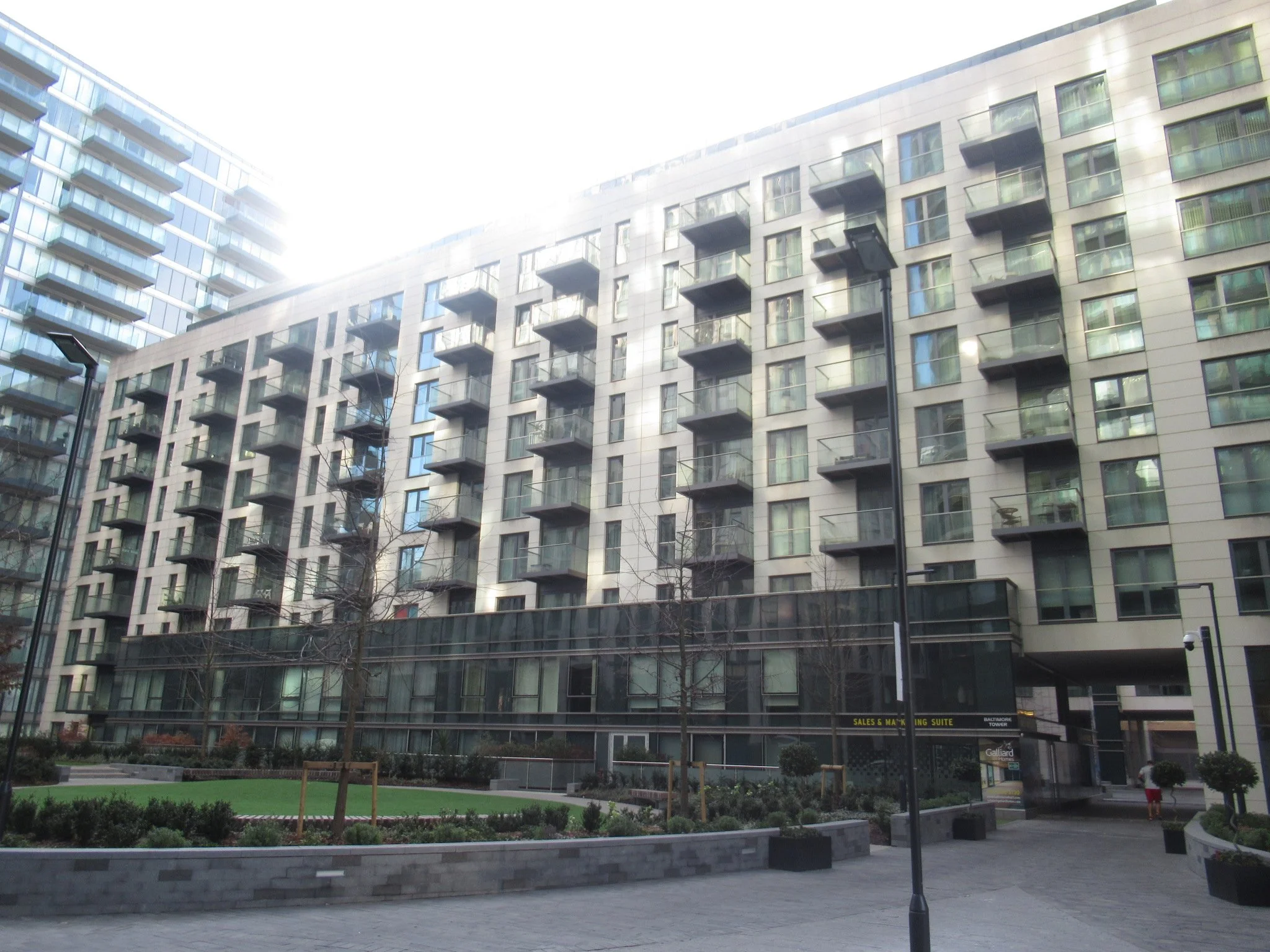 Modern residential building with multiple balconies and large windows, surrounding a landscaped courtyard with trees, benches, and pathways.