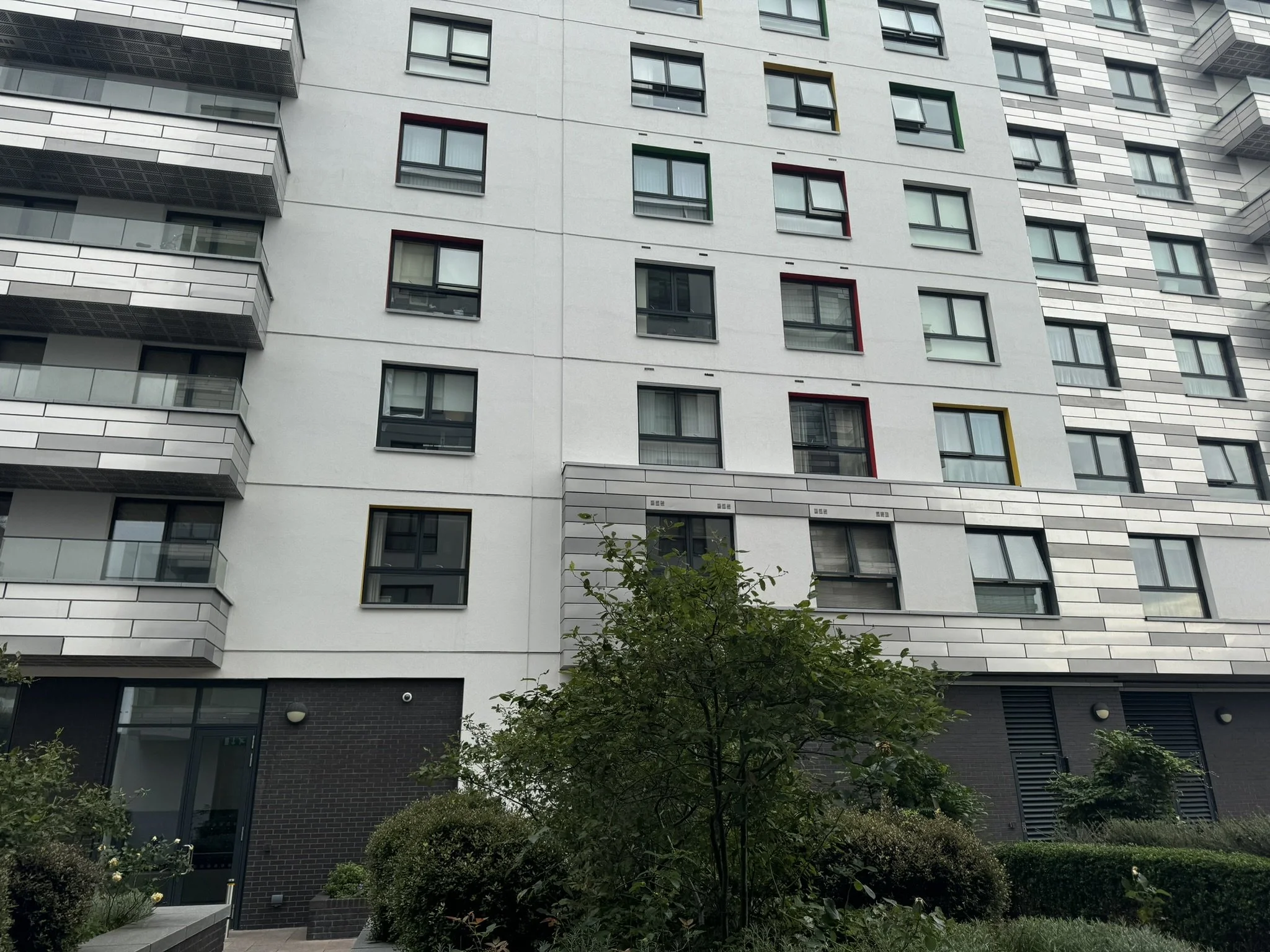 Modern multi-story apartment building with geometric window frames, balconies, and landscaped greenery in the foreground.