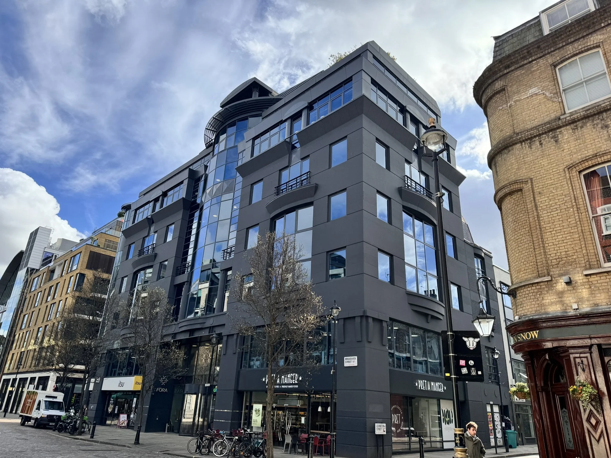 Modern dark gray multi-story building with large glass windows on a city street, surrounded by other buildings, trees, and street lamps, with a blue sky and clouds above.