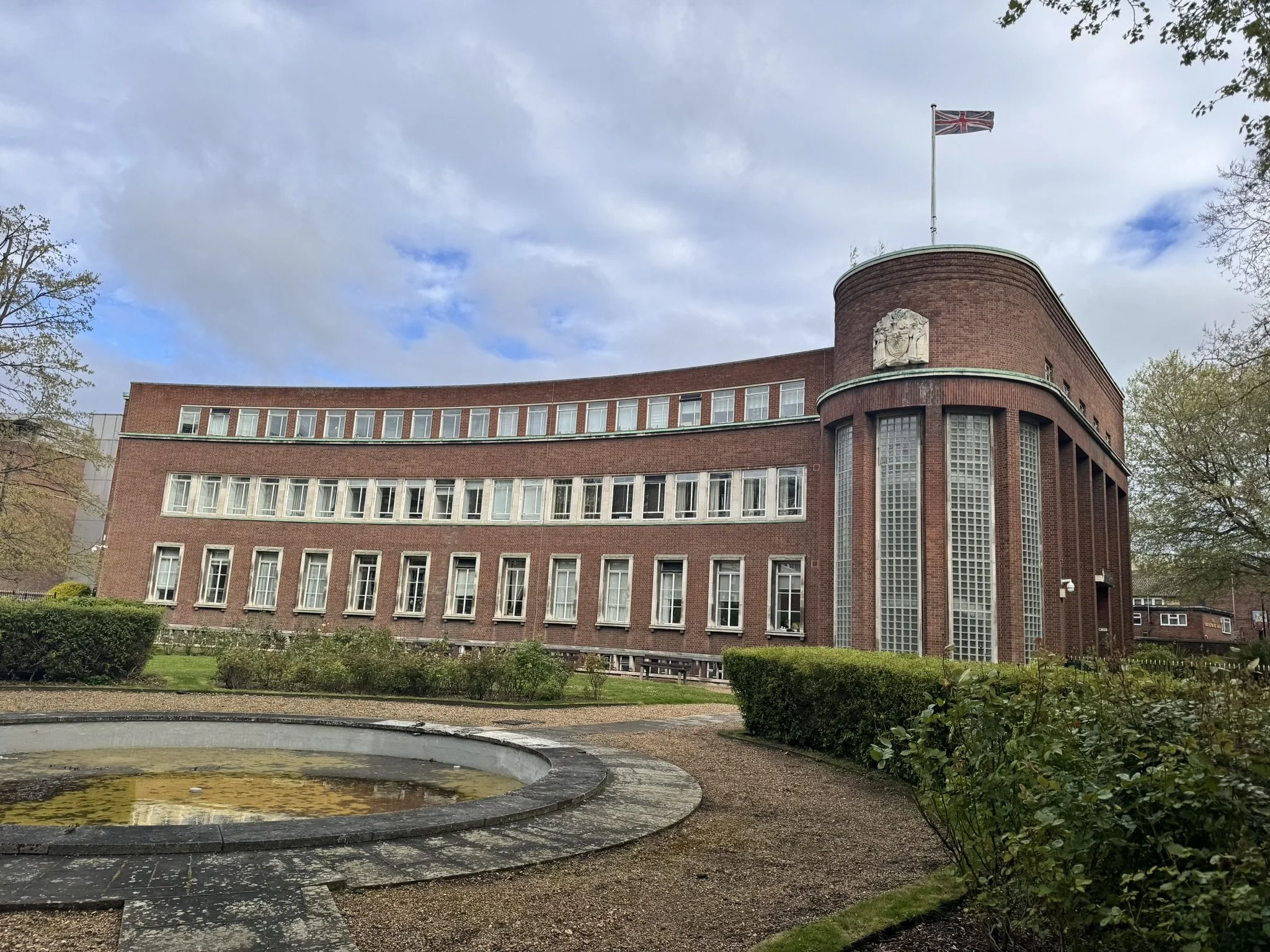A large, red brick building with curved architecture, multiple vertical windows, and a flagpole with the Union Jack flag on top. A small round pond is in the foreground, surrounded by a garden with bushes and trees. The sky is partly cloudy.