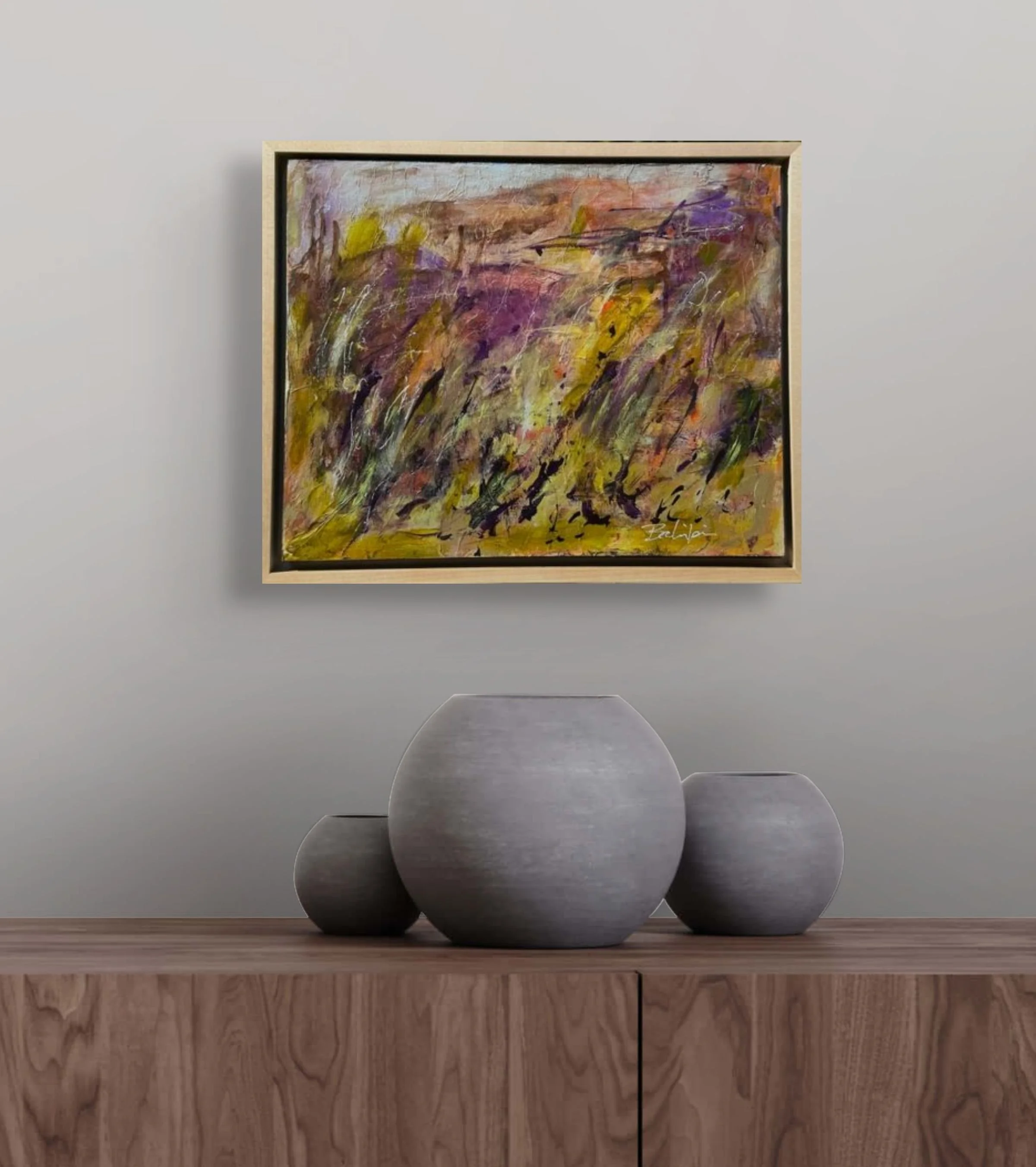 A still life arrangement featuring three round, textured ceramic vases in shades of gray and black, placed on a wooden surface in front of a framed abstract painting with colorful brushstrokes on a gray wall background.