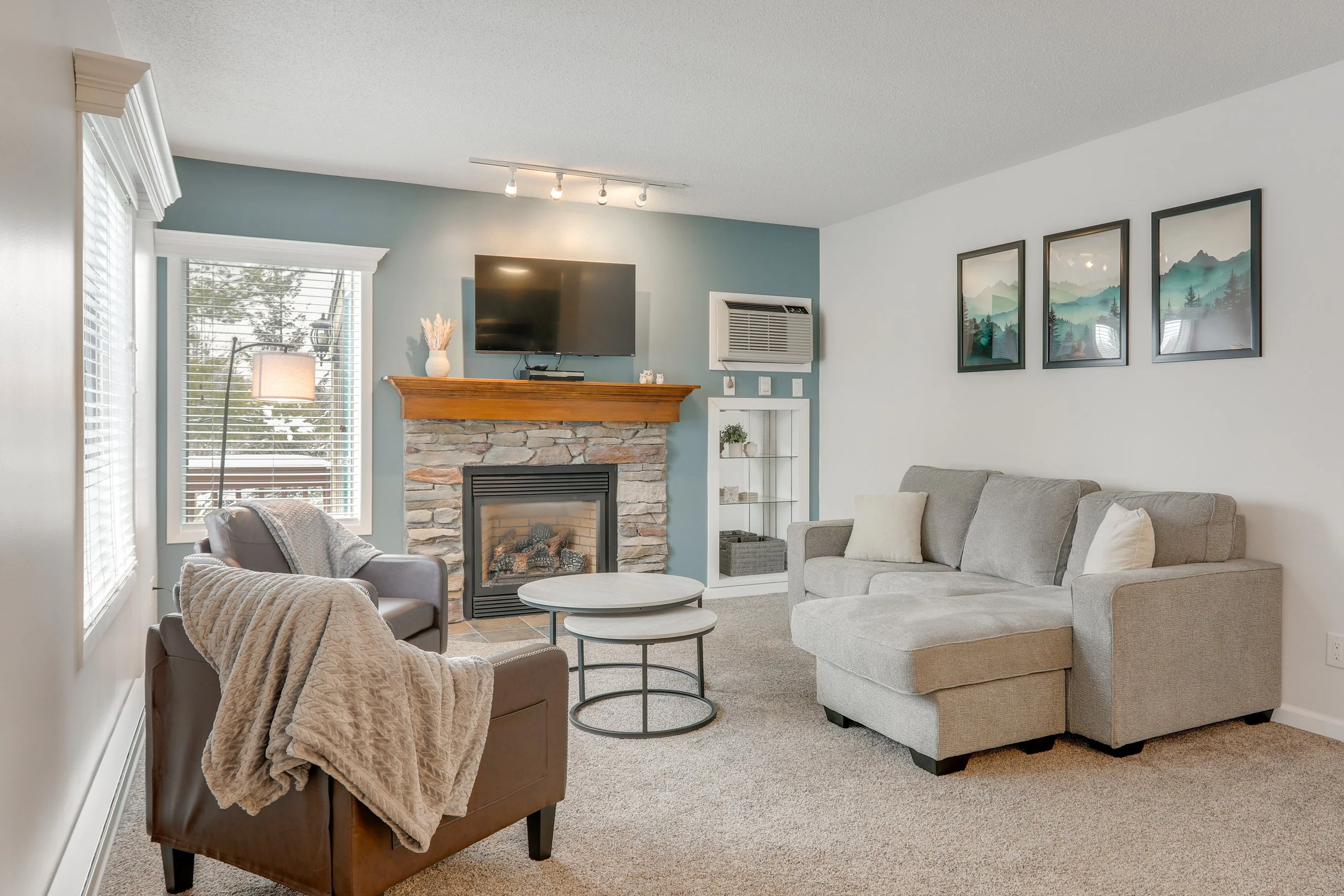 Living room with a gray sofa, a brown armchair with a blanket, a white and gray nested coffee table, a stone fireplace with a wooden mantle, a window with blinds, a floor lamp, wall art, and a mounted TV.