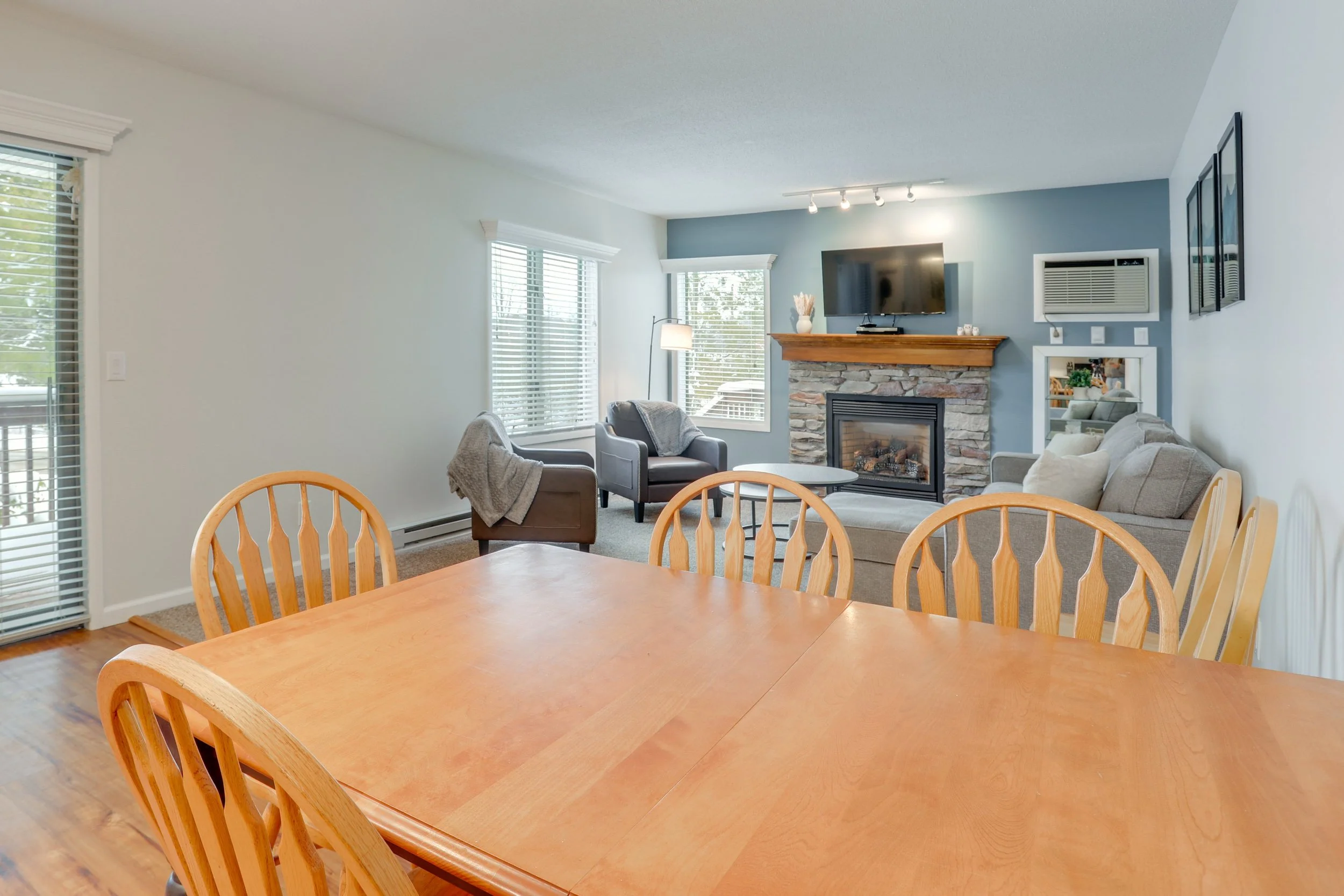 Open living room and dining area with wooden dining table, light wood chairs, gray sofa, patterned accent chair, stone fireplace with wooden mantle, wall-mounted TV, large windows with white blinds, and hardwood floors.