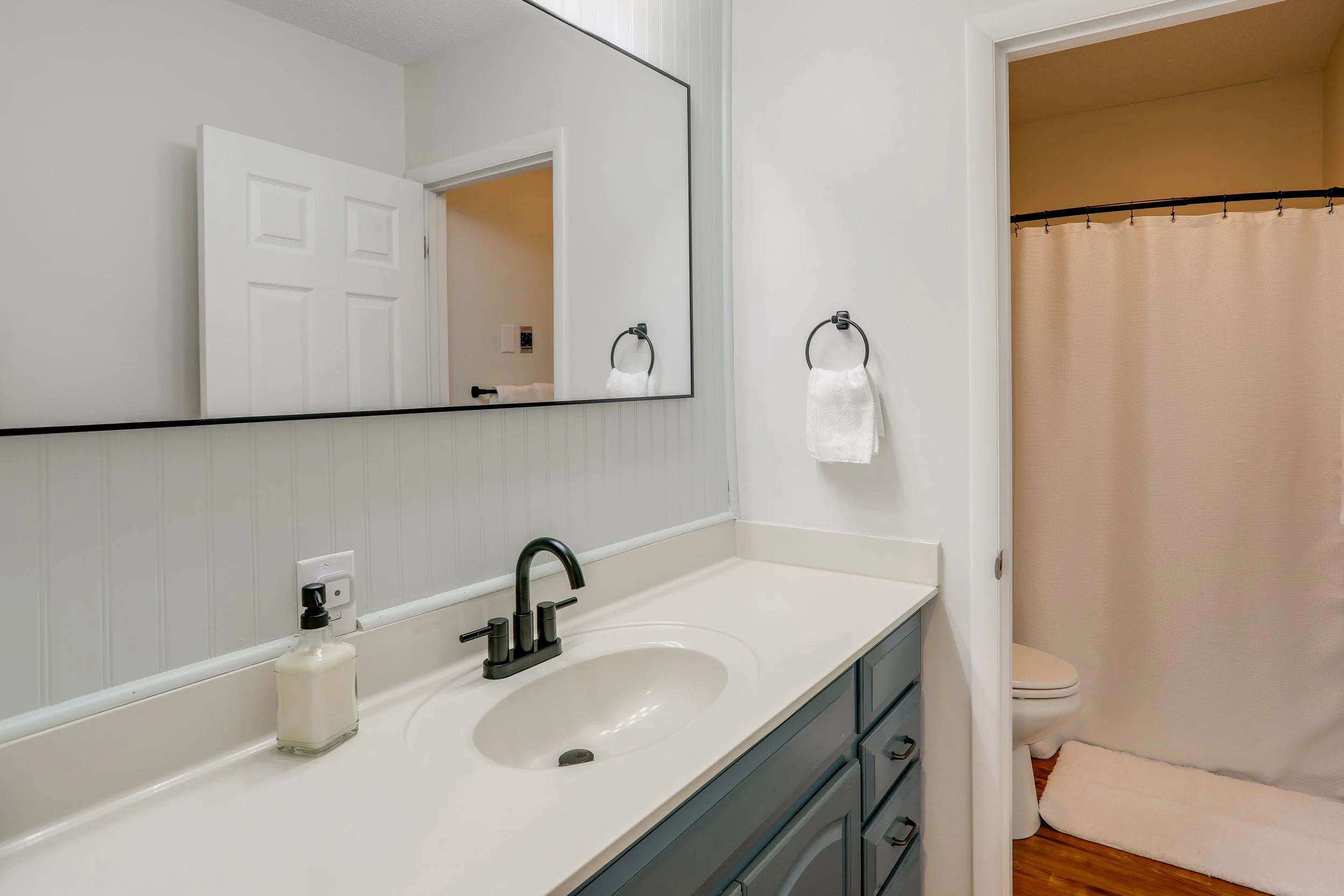 Bathroom with white vanity and mirror, black faucet, white countertop, soap dispenser, white towels on black ring, open door revealing a toilet with beige shower curtain and brown flooring.